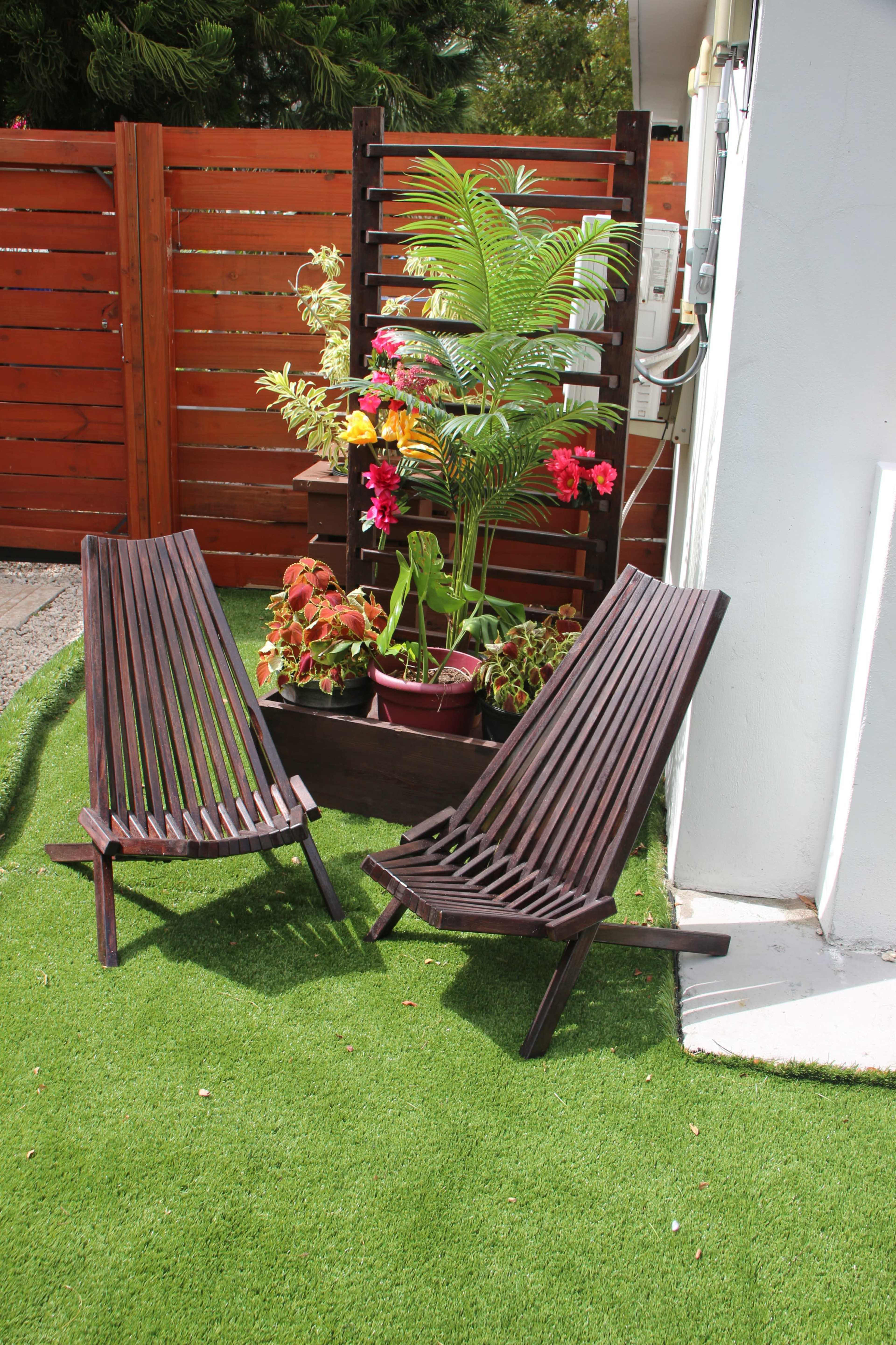 Two wooden lounge chairs are positioned on artificial grass next to a flower planter with various plants and bright flowers against a wooden fence.