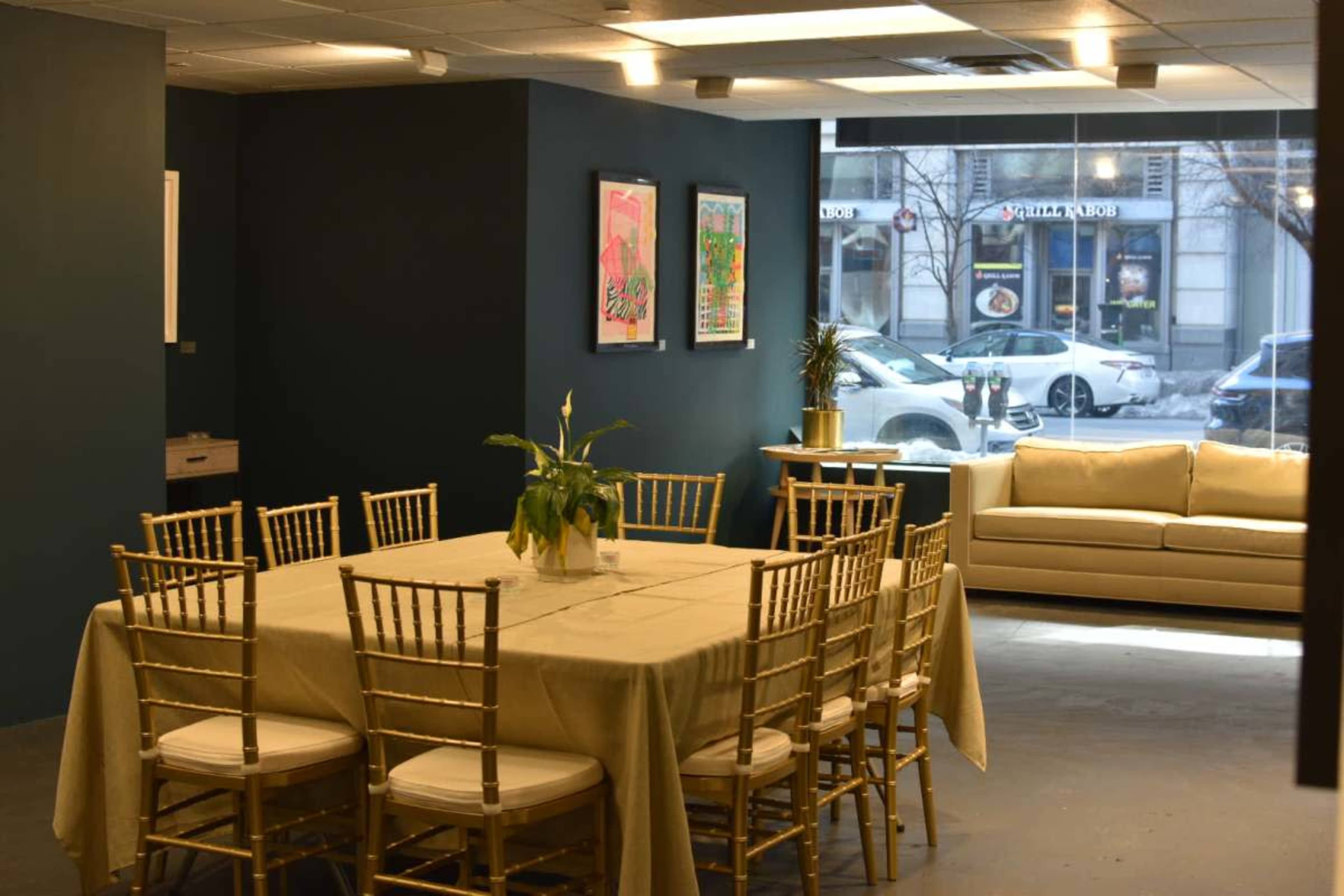 A large dining table with gold chairs is set in a room with dark blue walls, featuring artwork and a couch near a window overlooking a city street.