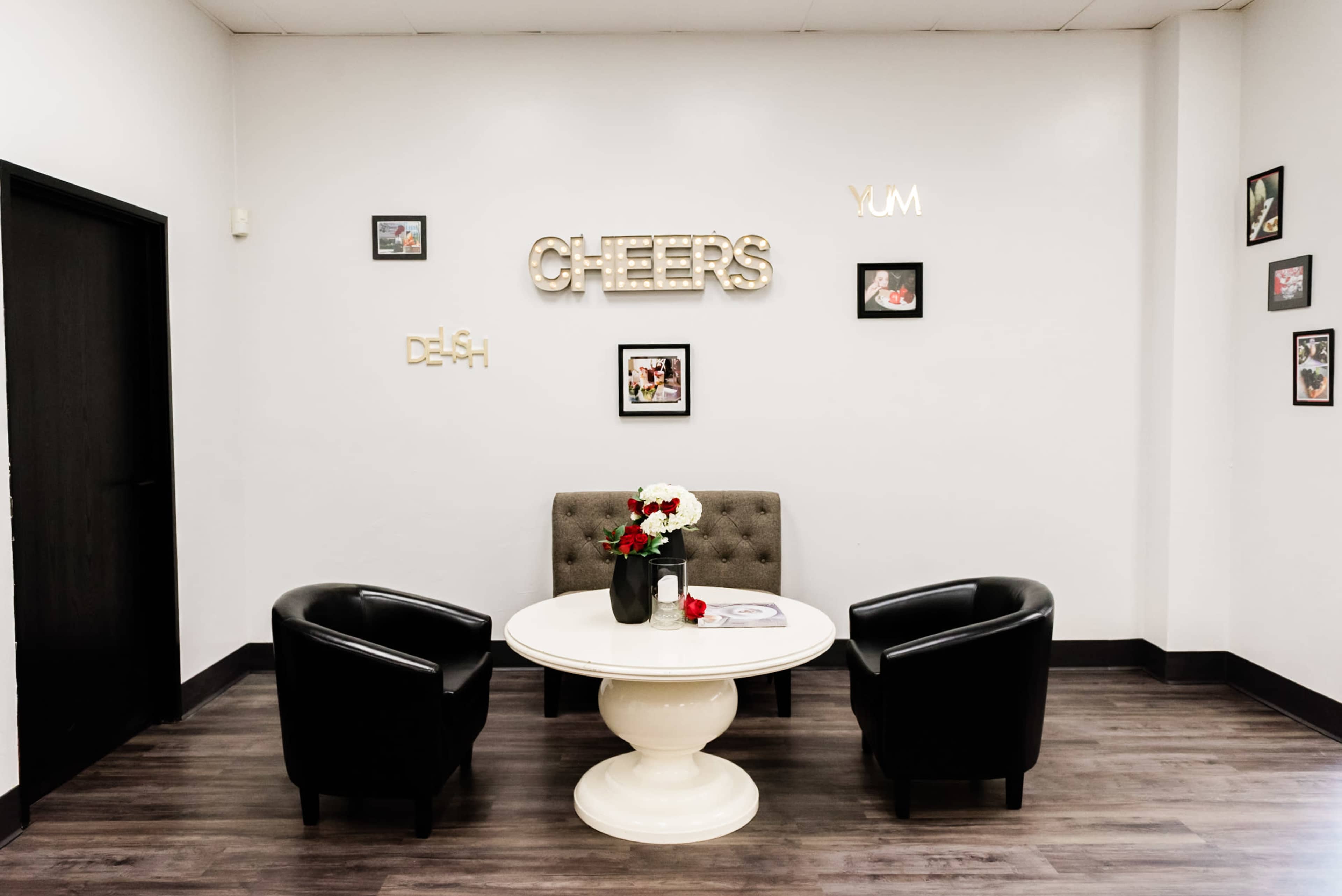 A small seating area features a round white table with a floral centerpiece, flanked by two black chairs, against a wall adorned with framed pictures and decorative letters spelling "CHEERS," "YUM," and "DISH."