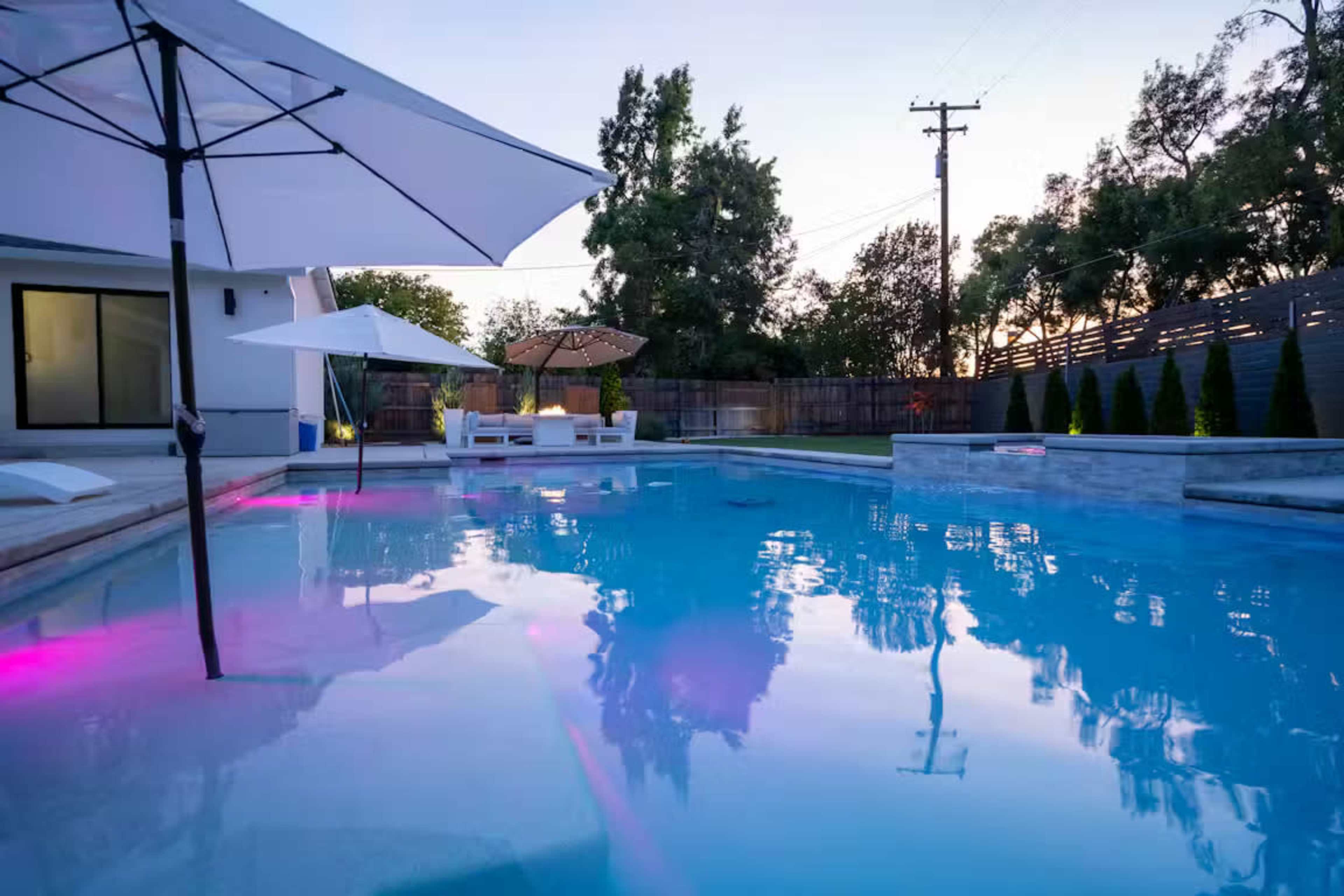 A serene backyard scene features a swimming pool with illuminated water, surrounded by lounge chairs and umbrellas under a clear sky.