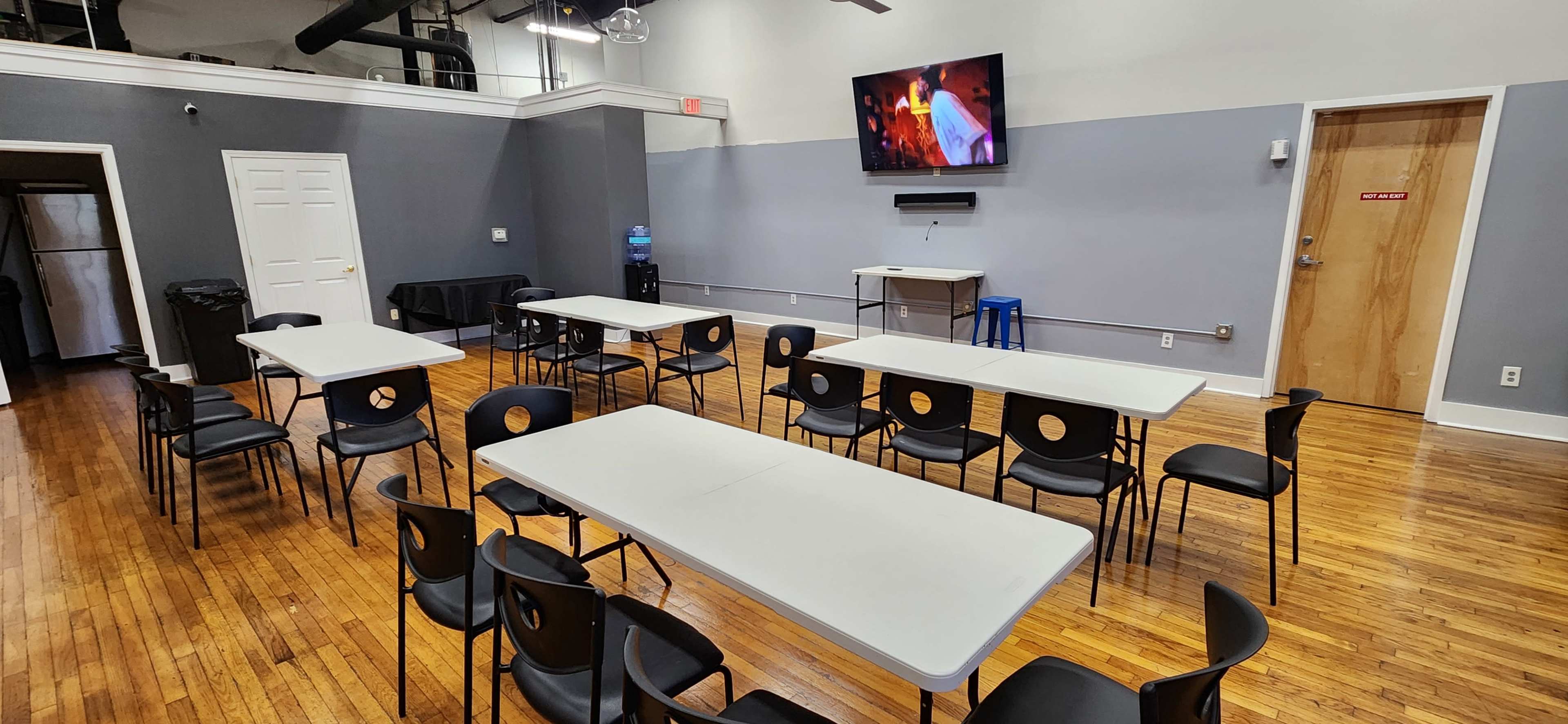 The image shows a set of tables and chairs arranged in a room with wood flooring, a television mounted on the wall, and a water dispenser in the corner.