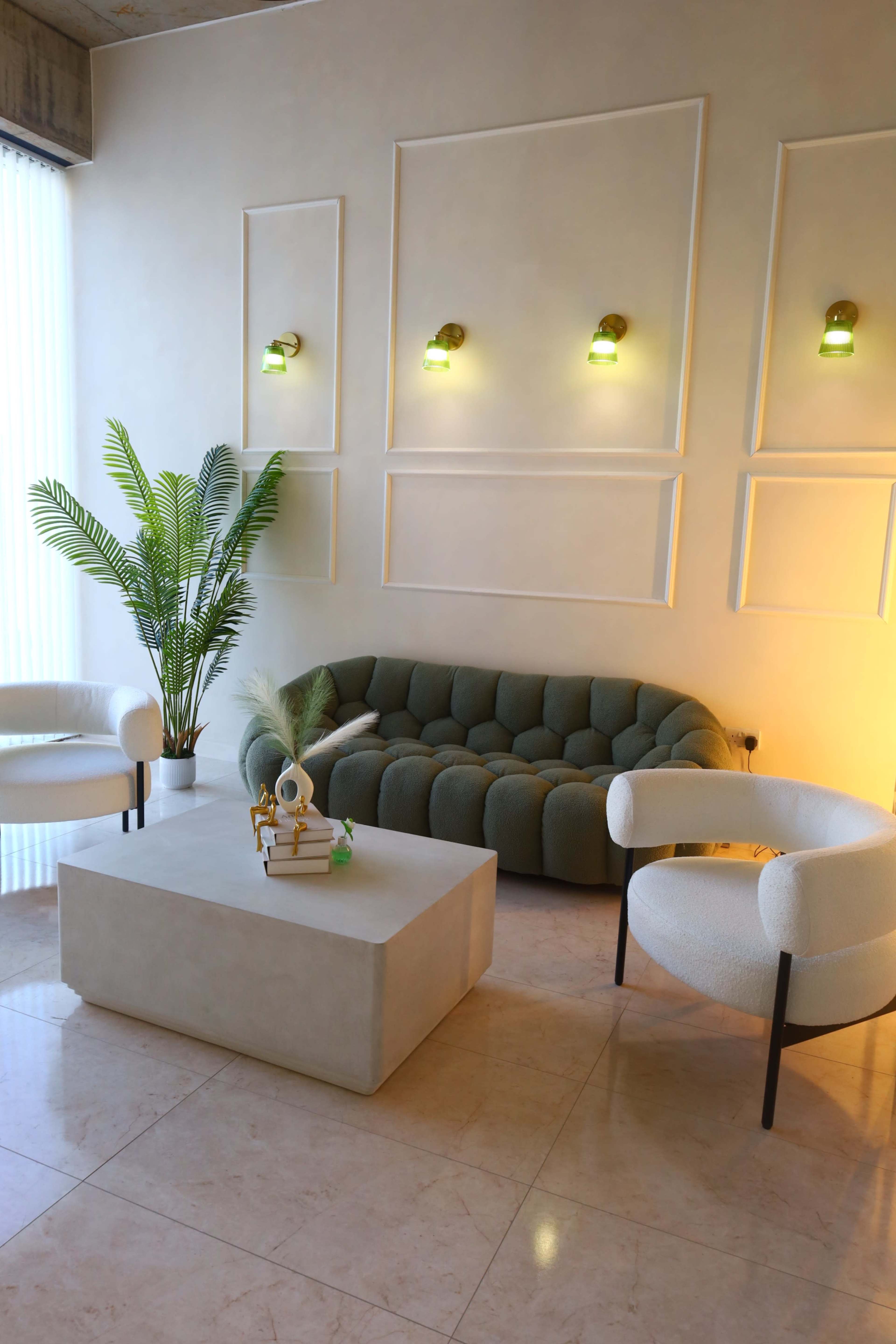The image shows a modern lounge area with a green sofa, two white armchairs, a low coffee table, and a potted plant against a light-colored wall with wall sconces.