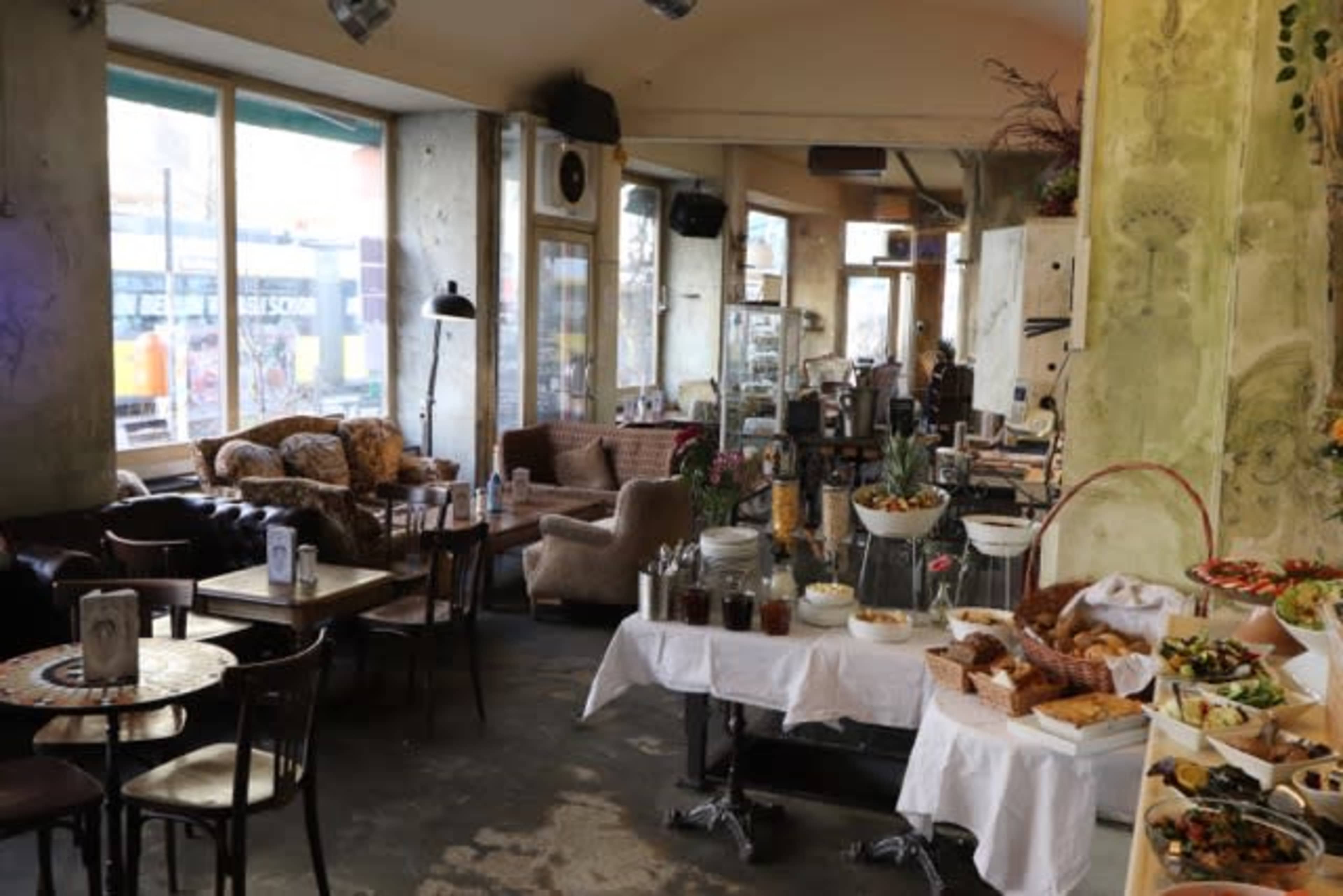 The image shows a cozy café interior with a buffet table featuring various foods and a seating area with chairs and couches.
