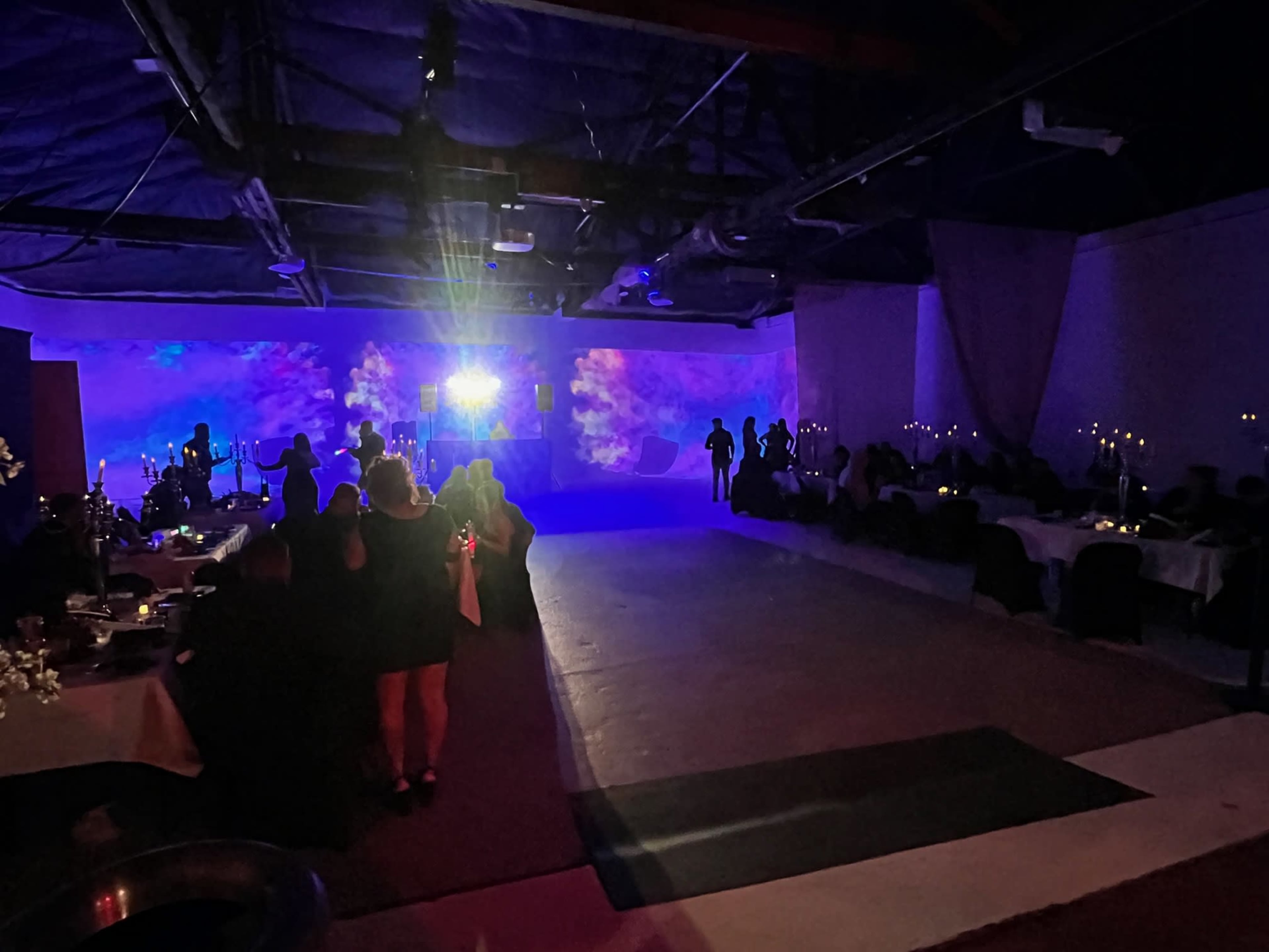 A large event space is filled with tables and chairs, featuring a band in the background and colorful lighting casting patterns on the walls.