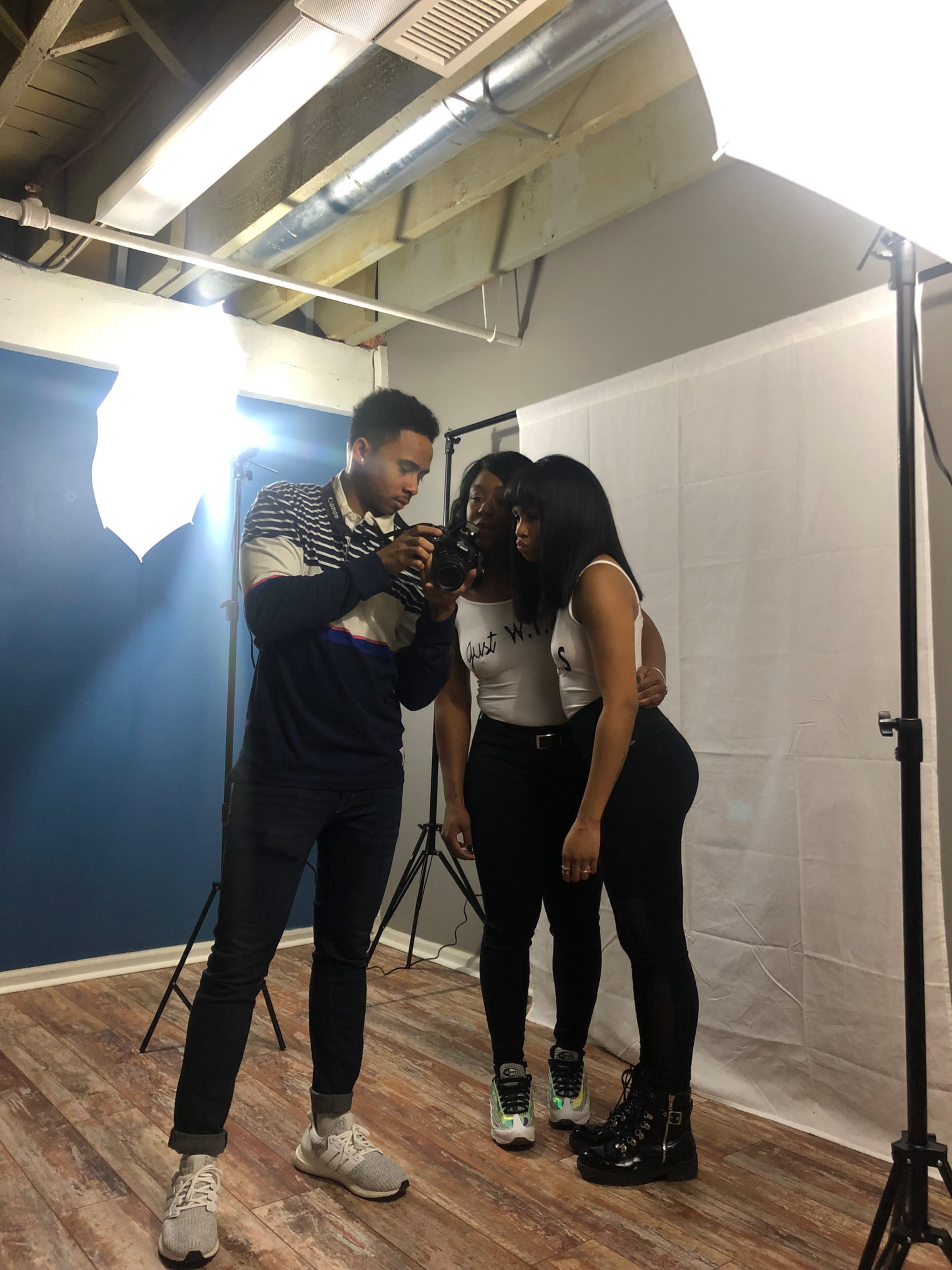 A photographer adjusts his camera while two models pose for a photoshoot in a studio setting.