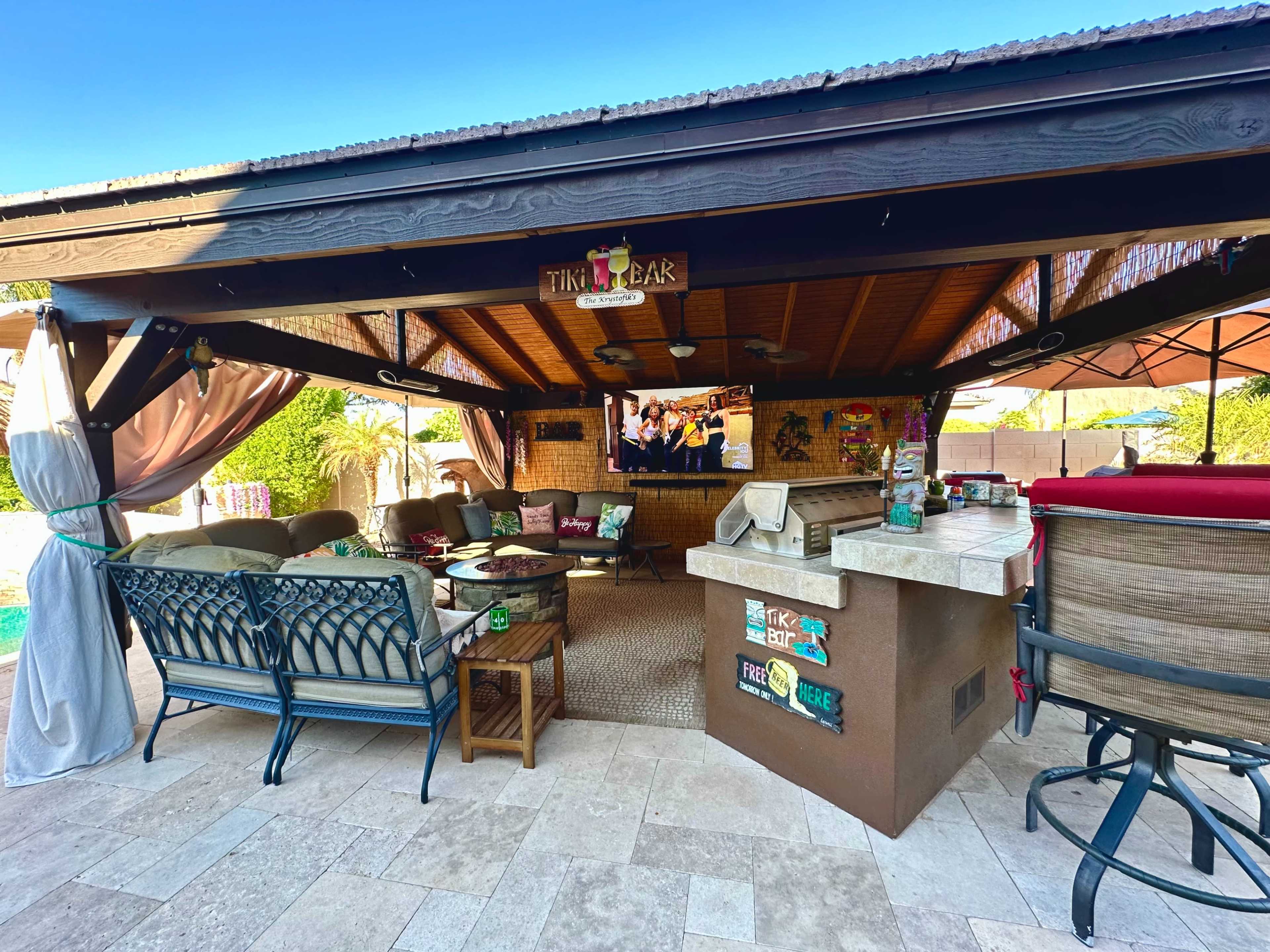 A covered outdoor tiki bar area with seating, a television mounted on the wall, and a grill.