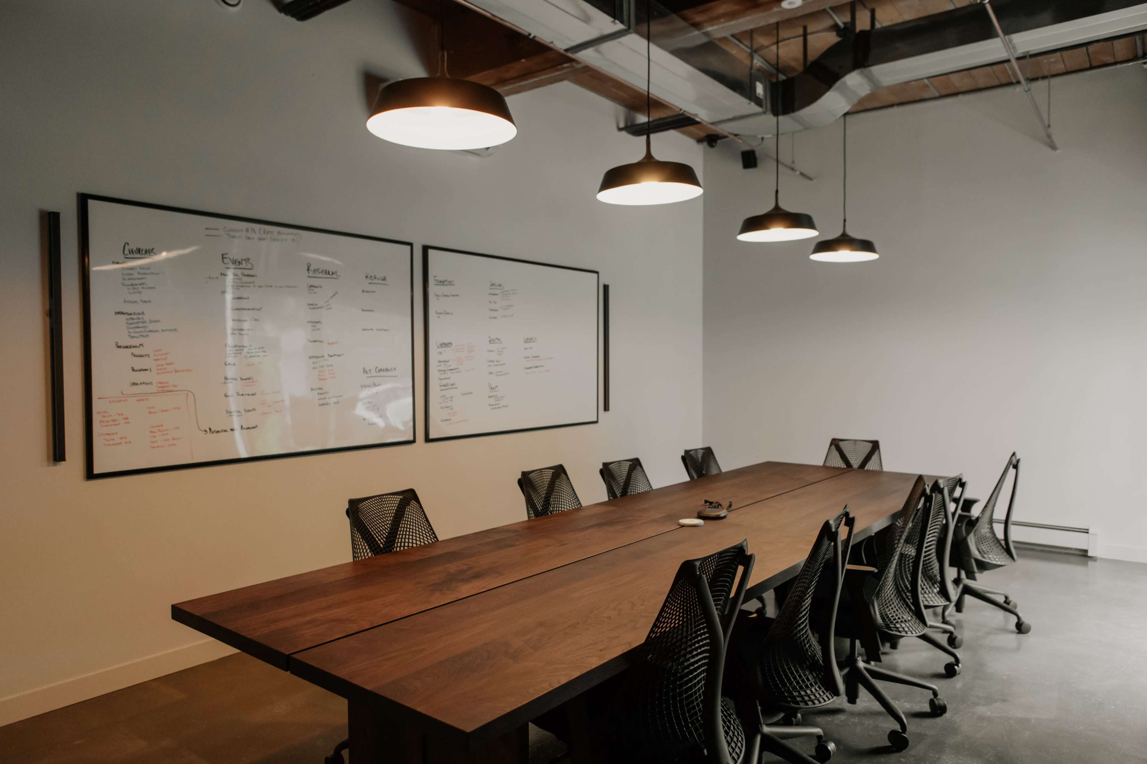 A conference room features a long wooden table surrounded by black mesh chairs and two large whiteboards mounted on the wall.
