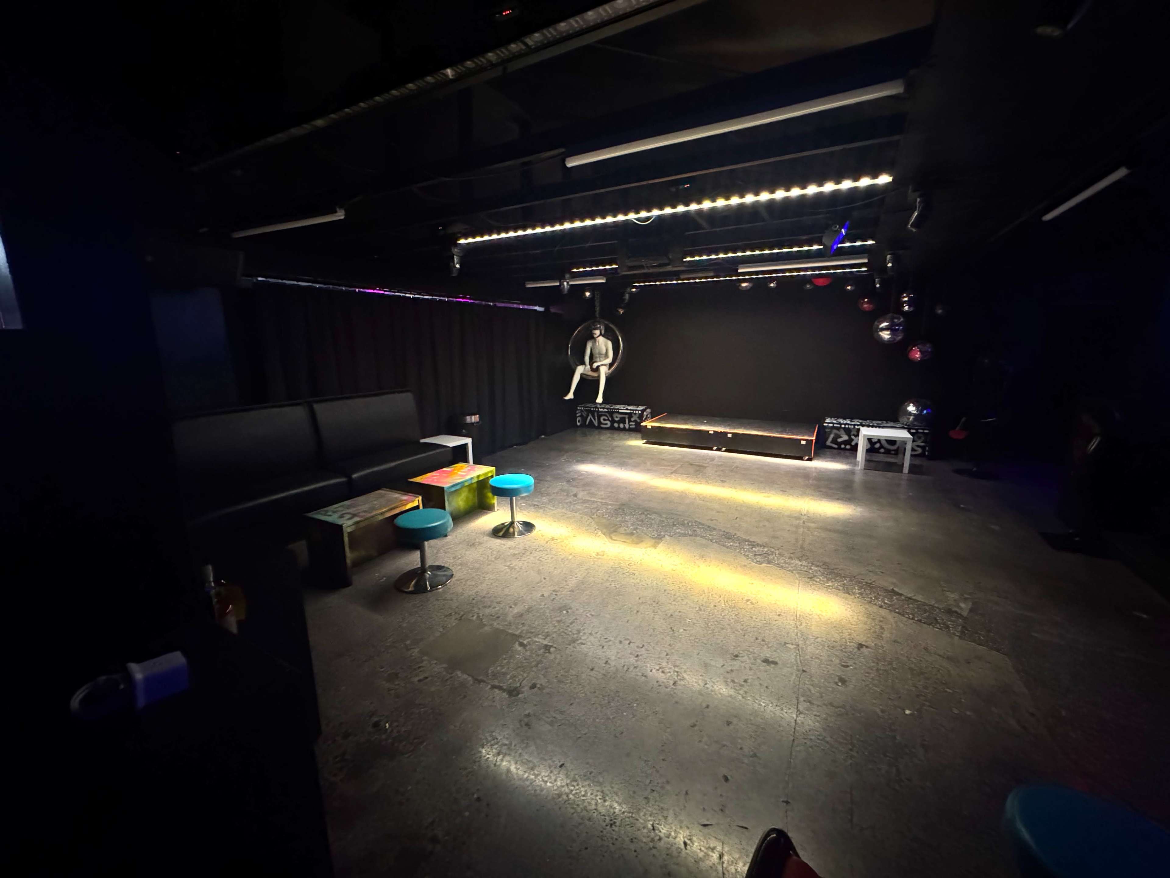 The image shows a dimly lit performance space with black walls, scattered seating, and a small stage equipped with lights and a disco ball.