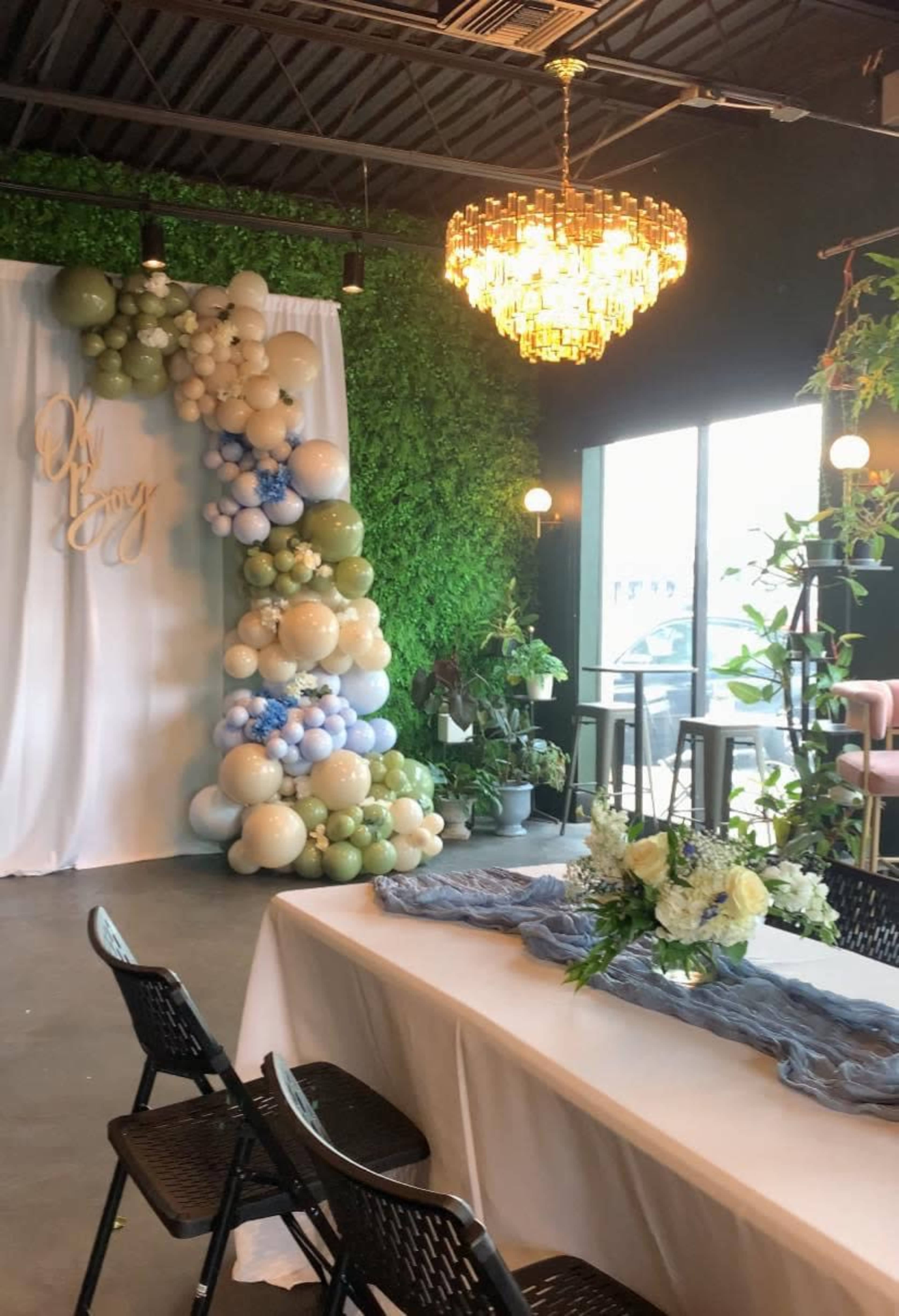 The image shows a decorated event space featuring a floral arrangement on a table, a backdrop with balloon decorations, and a chandelier hanging from the ceiling.
