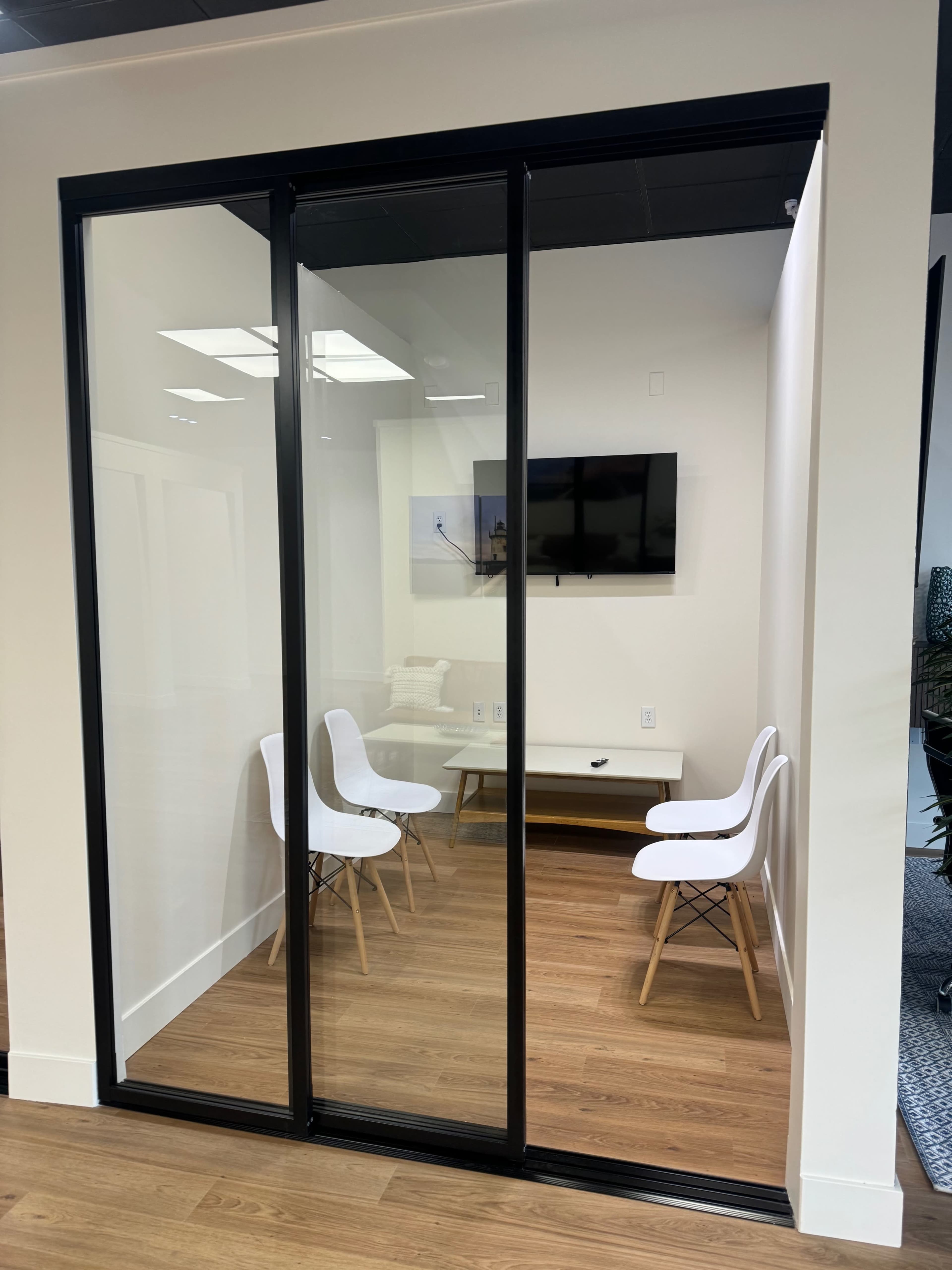A small meeting room with glass walls, containing a table and four white chairs, and a wall-mounted television.