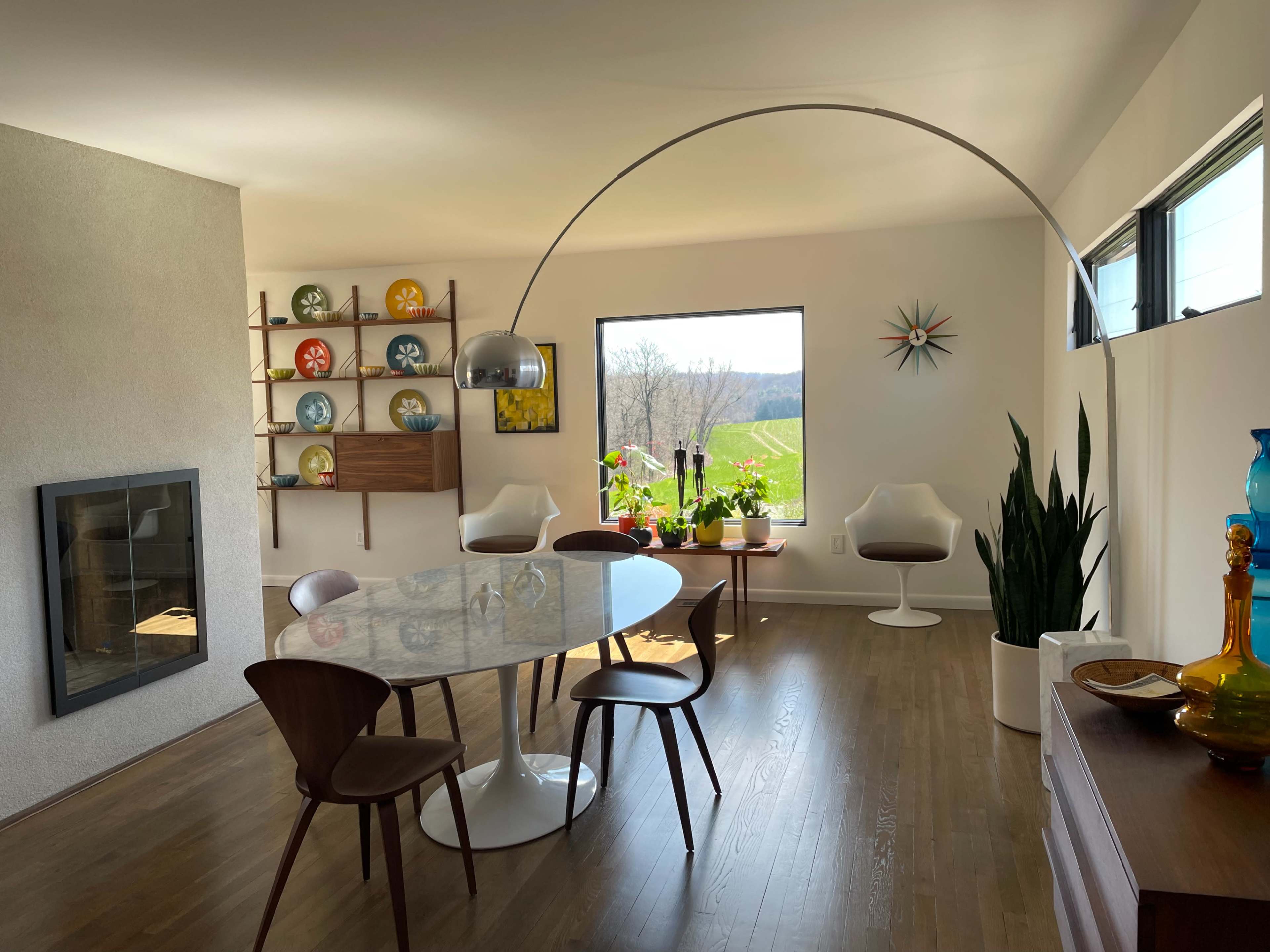 A modern dining room features a circular table surrounded by wooden chairs, a large window with a view of greenery, and a decorative shelf against the wall.