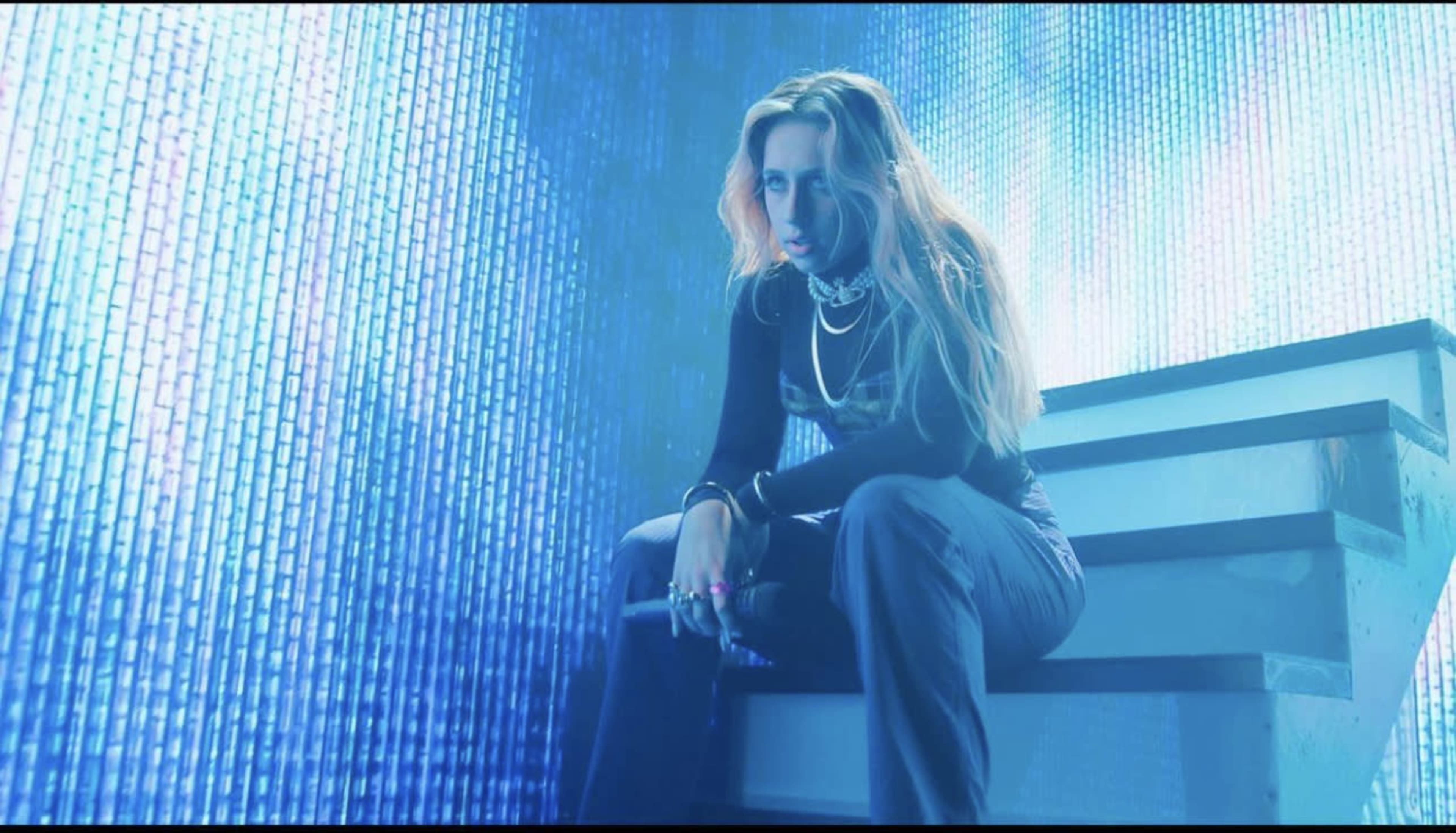 A performer sits on a staircase under vibrant blue lighting with a digital backdrop.