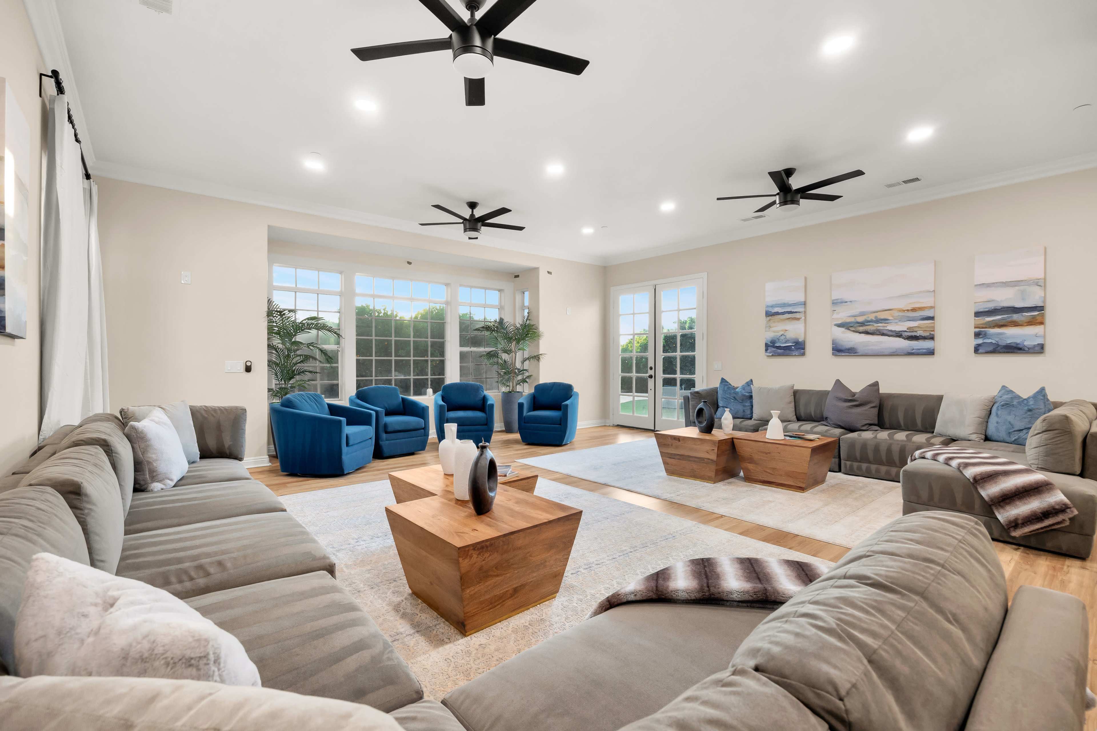 The spacious living room features two large seating areas with gray sofas and blue armchairs, complemented by wooden coffee tables and large windows.