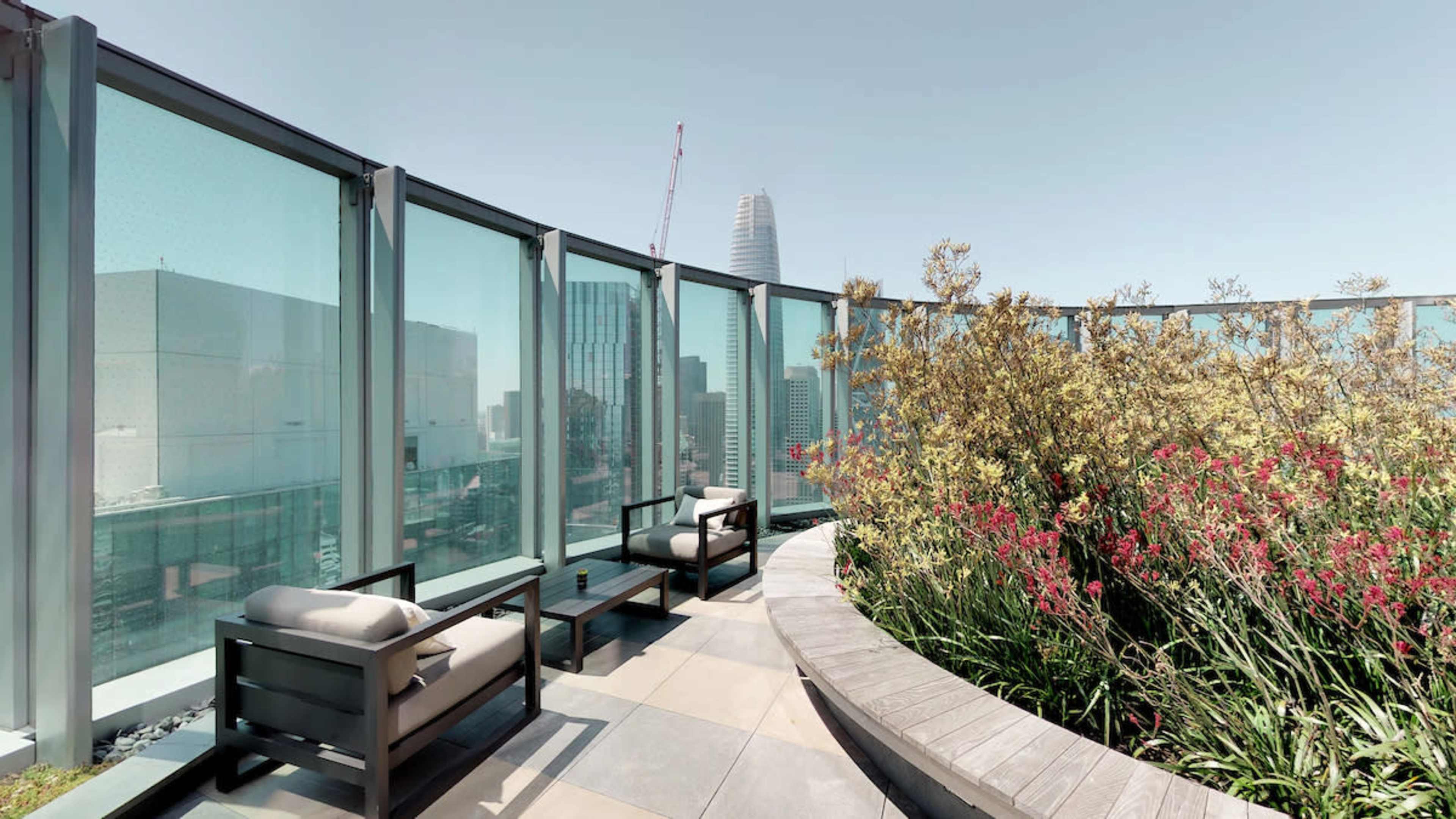 A rooftop terrace features modern seating arrangements surrounded by glass panels and plants, with a skyline view in the background.
