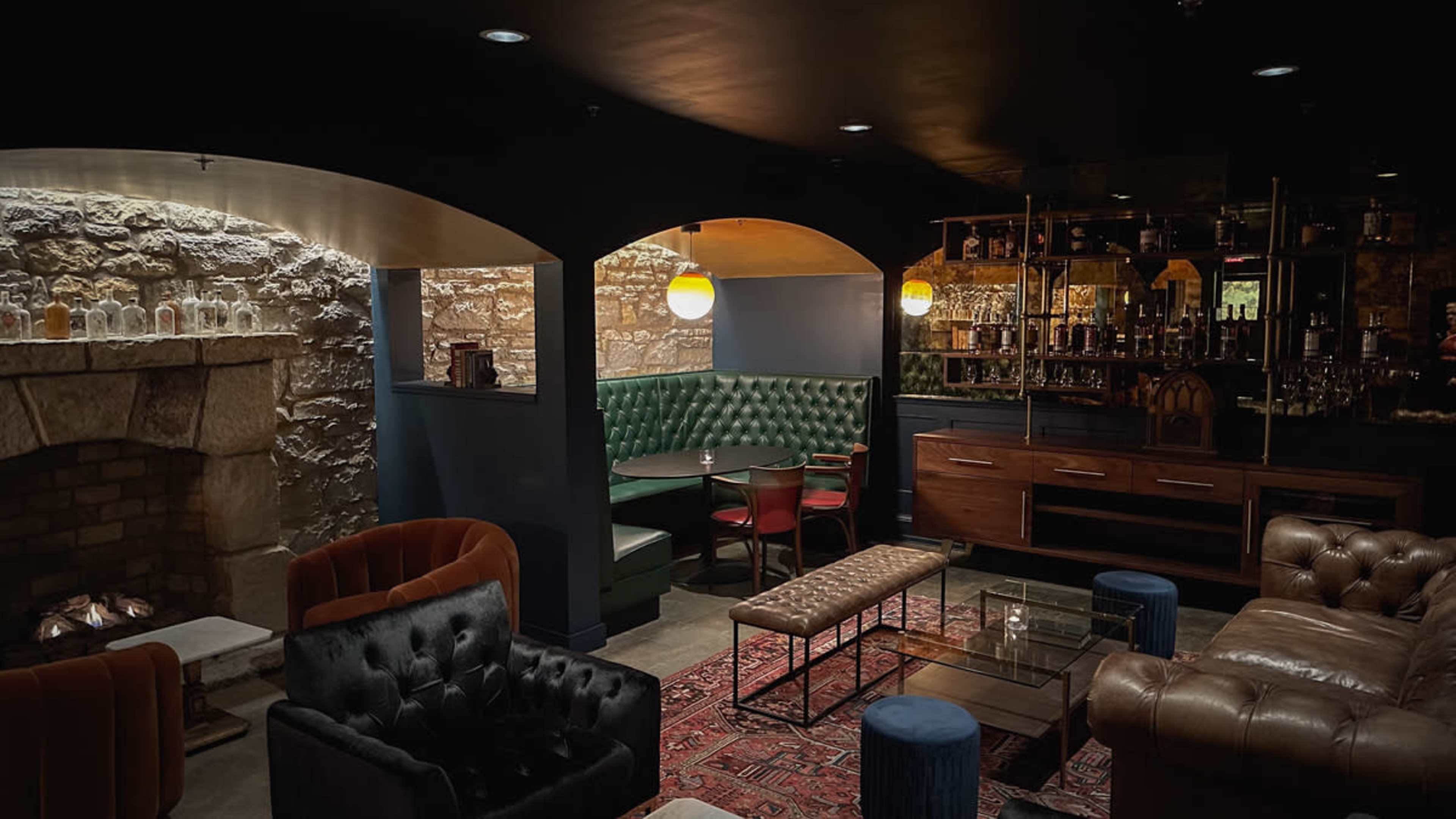 A dimly lit lounge area featuring plush seating, a stone wall with a fireplace, and a well-stocked bar in the background.