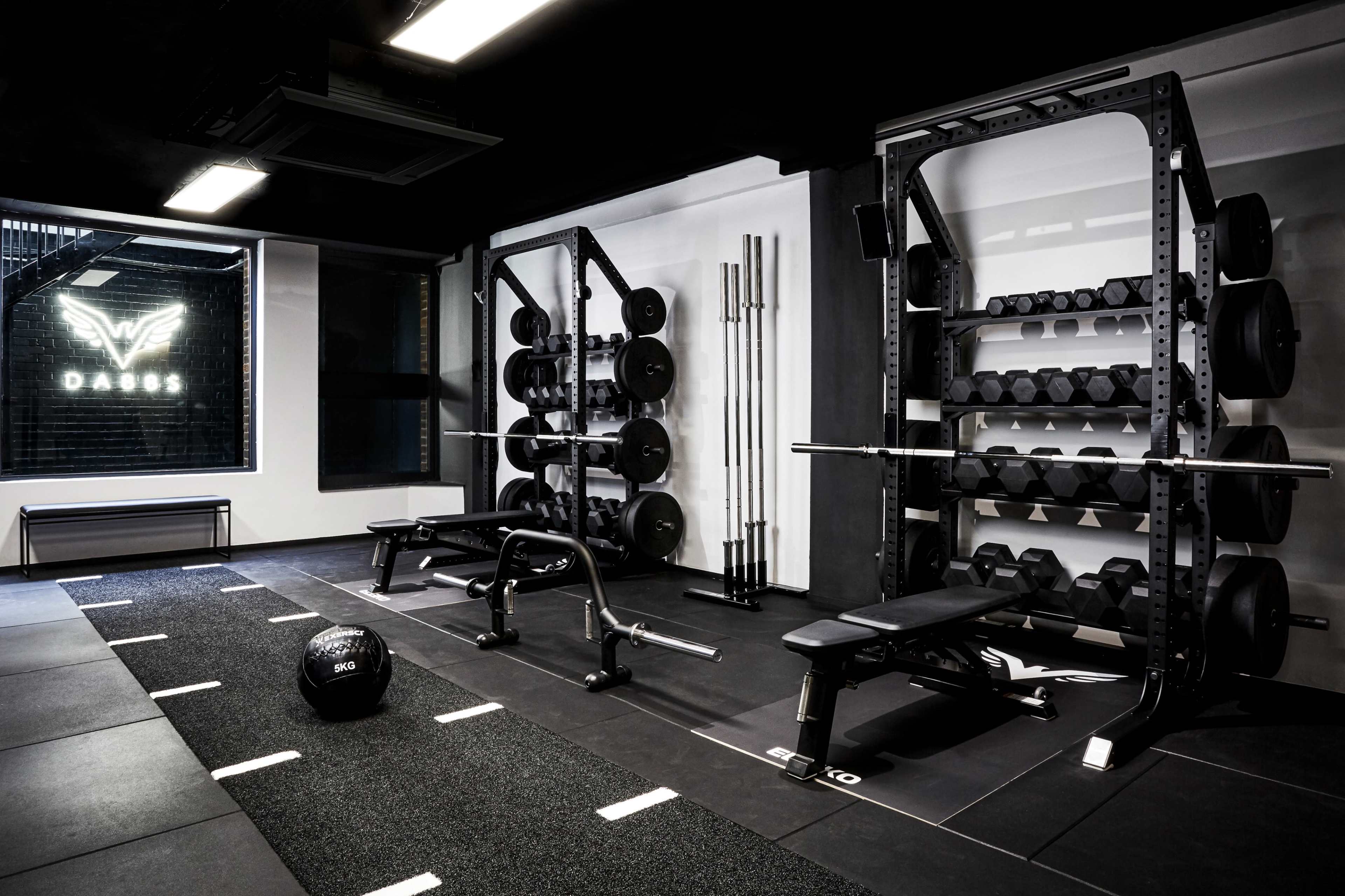 A modern gym interior featuring weightlifting equipment, including benches, dumbbells, and a barbell rack, set against a backdrop with a logo.