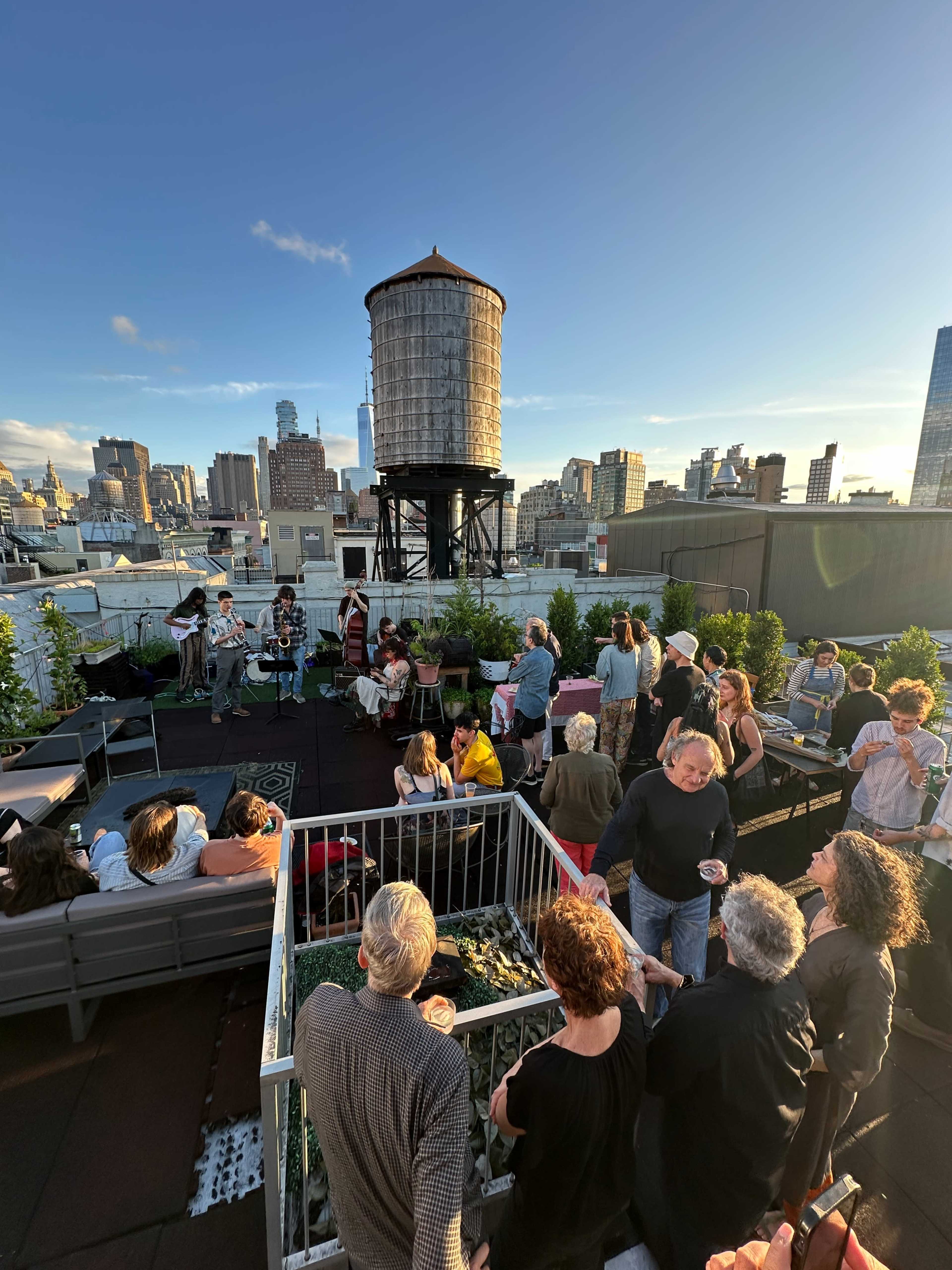 A crowd gathers on a rooftop terrace in a city for a live music performance as skyscrapers and a water tower stand in the background.