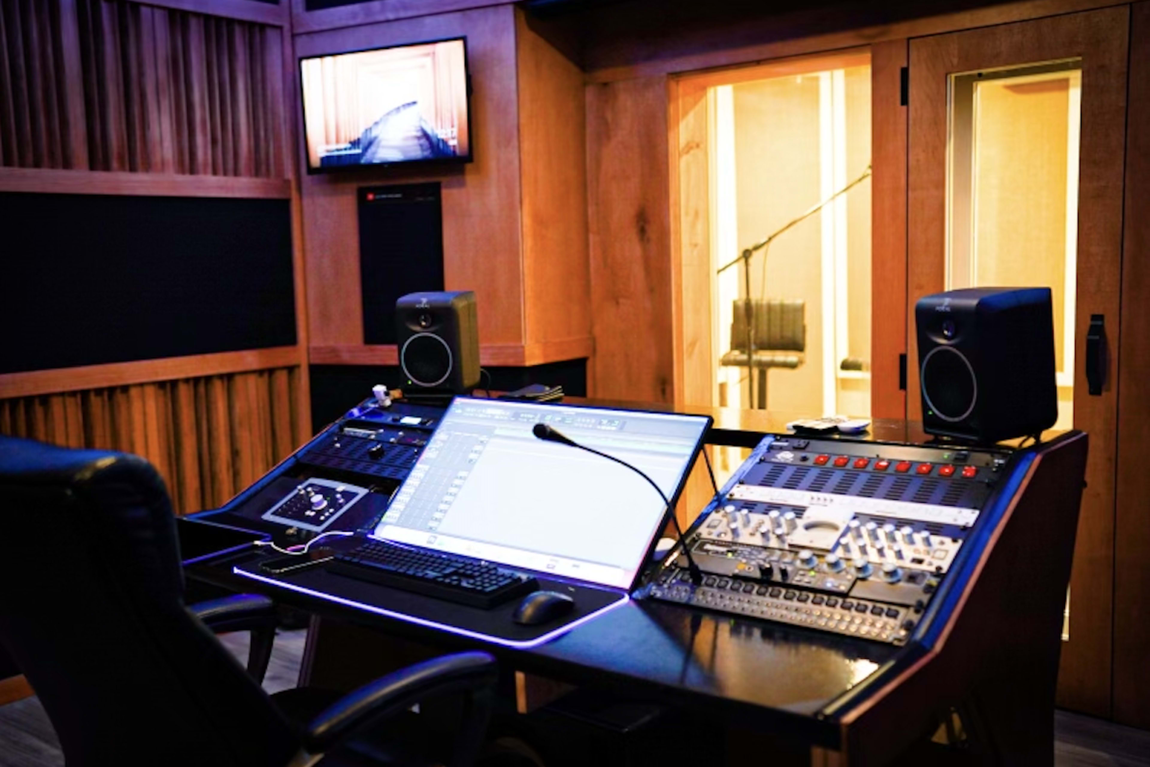 A sound recording studio features a control desk with audio mixing equipment, a computer monitor, and speakers, alongside a glass-enclosed recording booth.