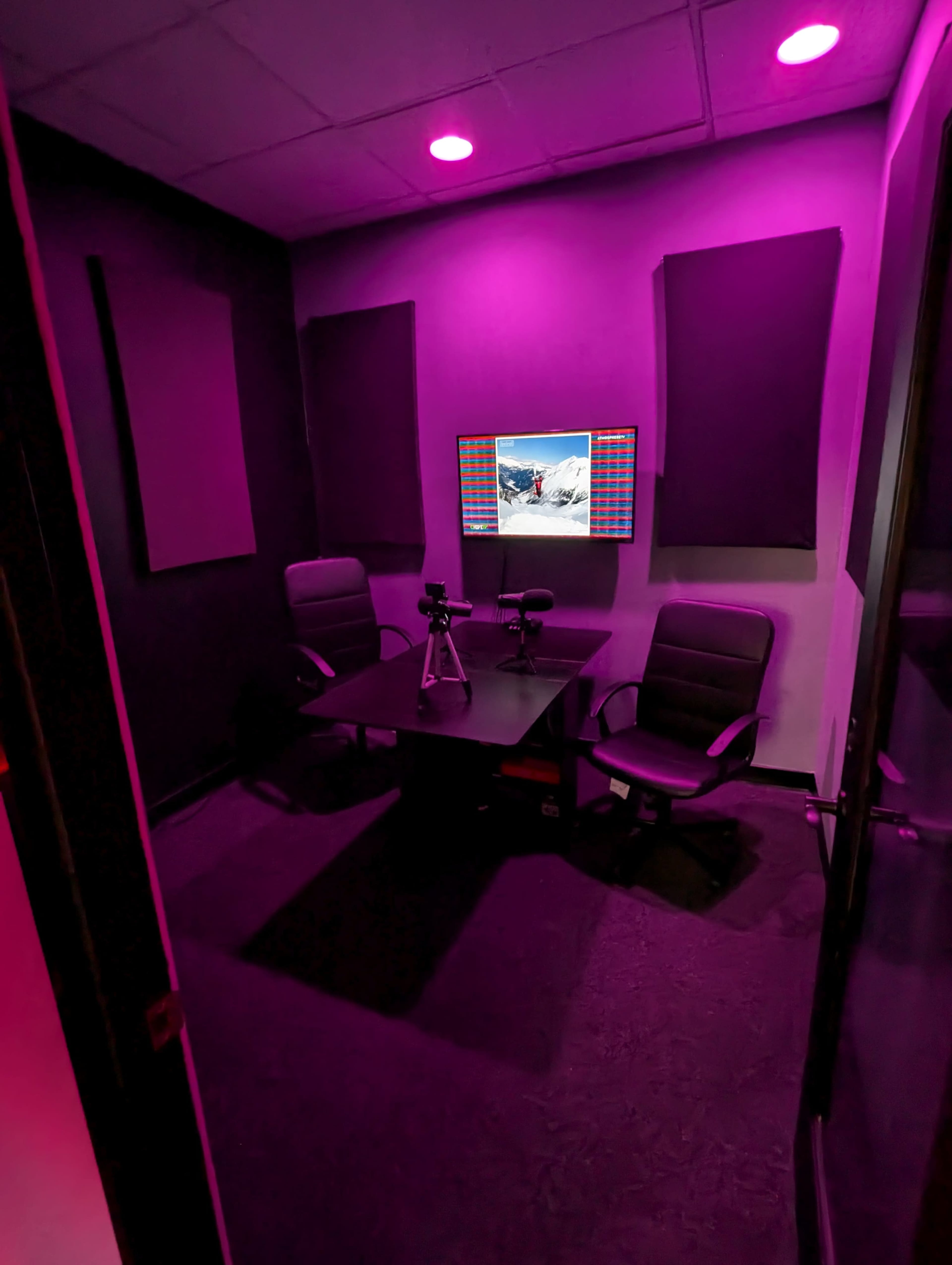 A small conference room with two chairs, a table, and a mounted screen, illuminated by purple lighting.