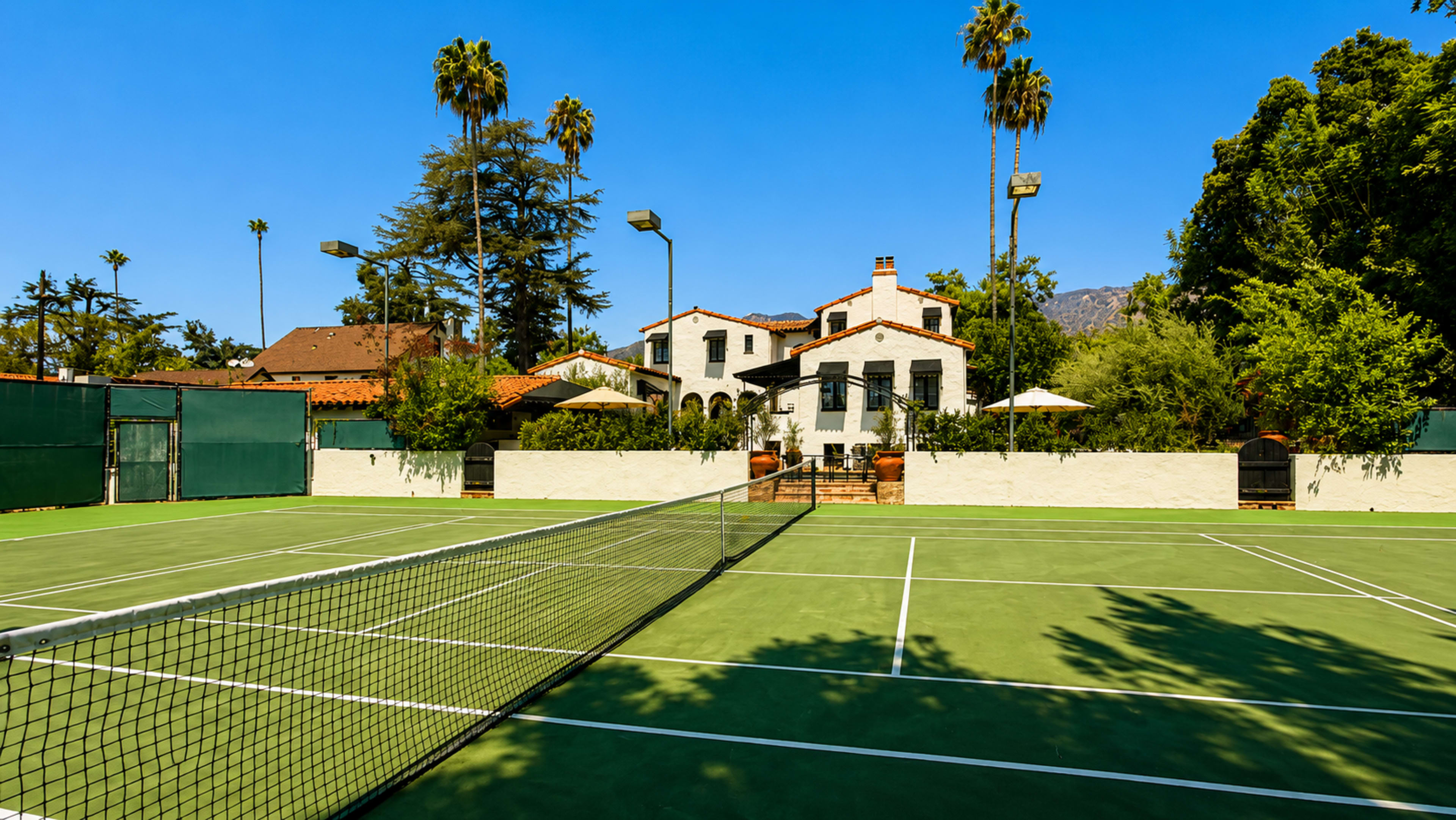 The image shows a tennis court in front of a large house surrounded by tall palm trees and other greenery.