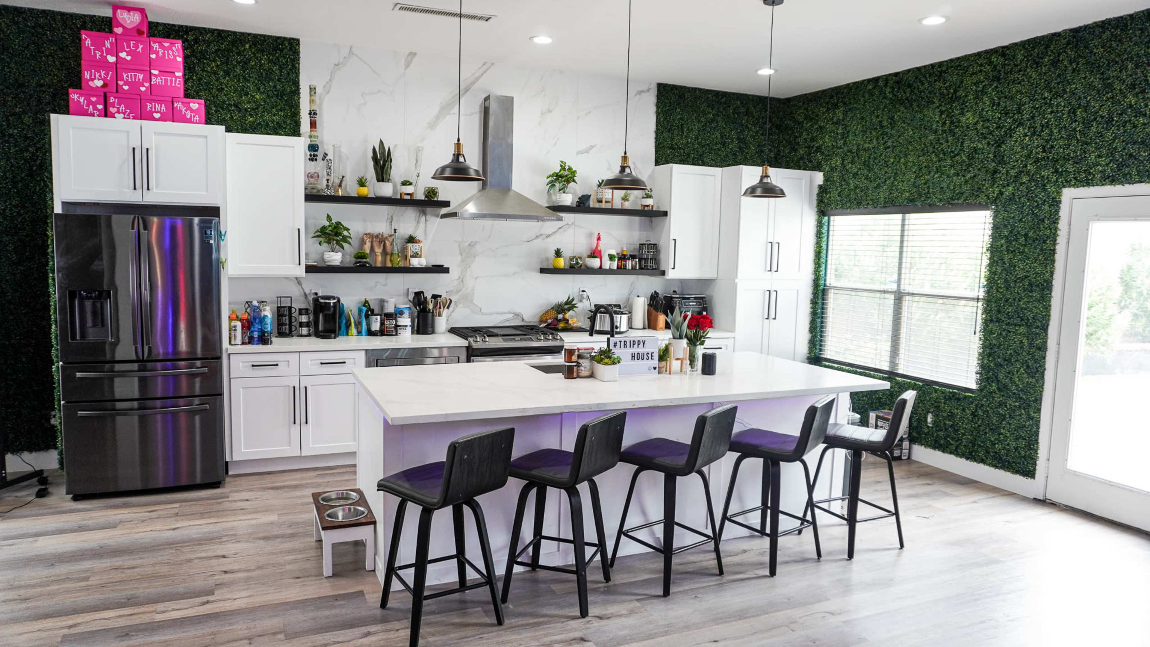 A modern kitchen with white cabinets, a large island with seating, and a decorative green wall featuring various plants.