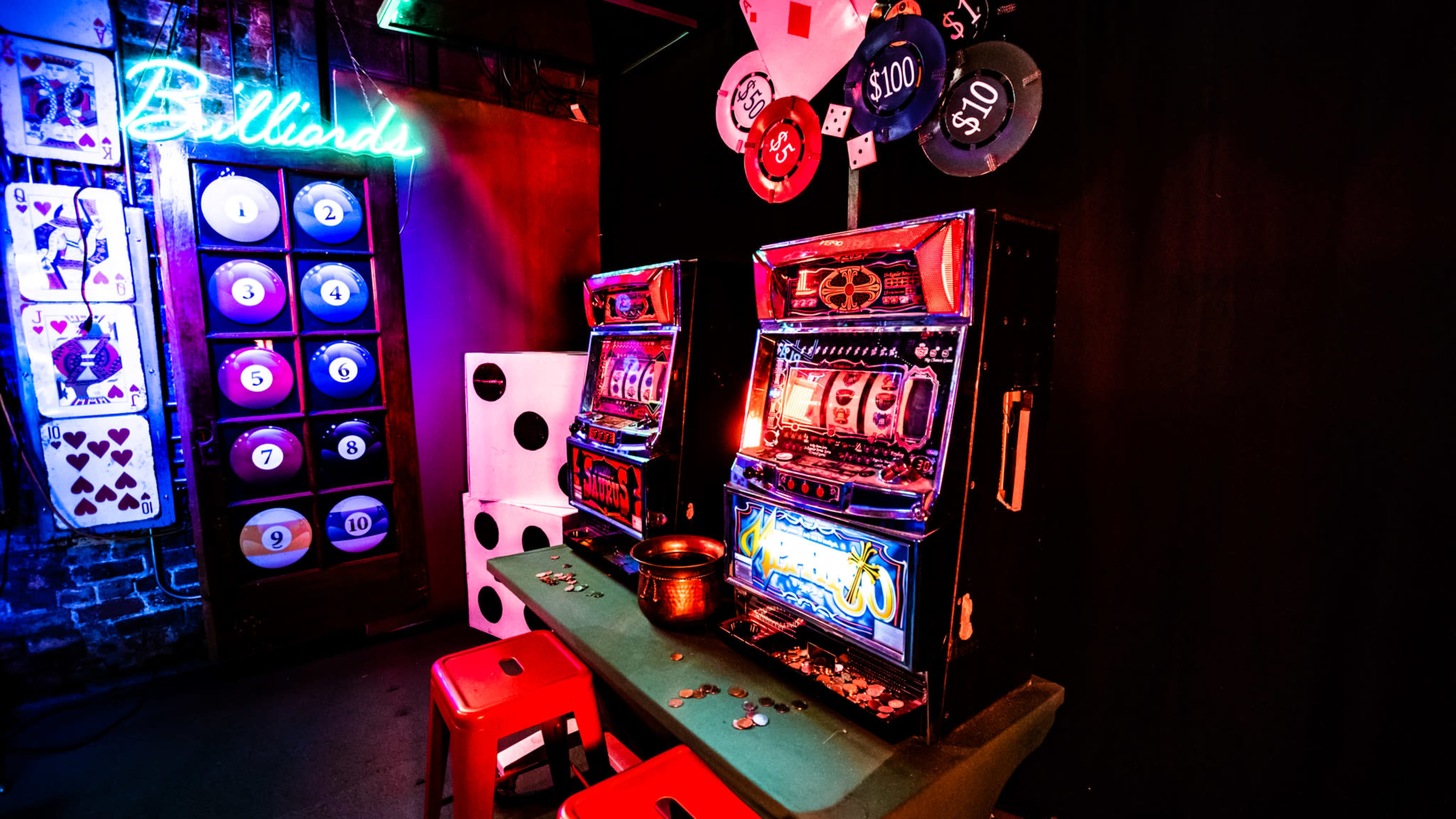 Two vintage slot machines illuminated by neon lights in a dimly lit room, accompanied by playing cards and casino chips on a green table.