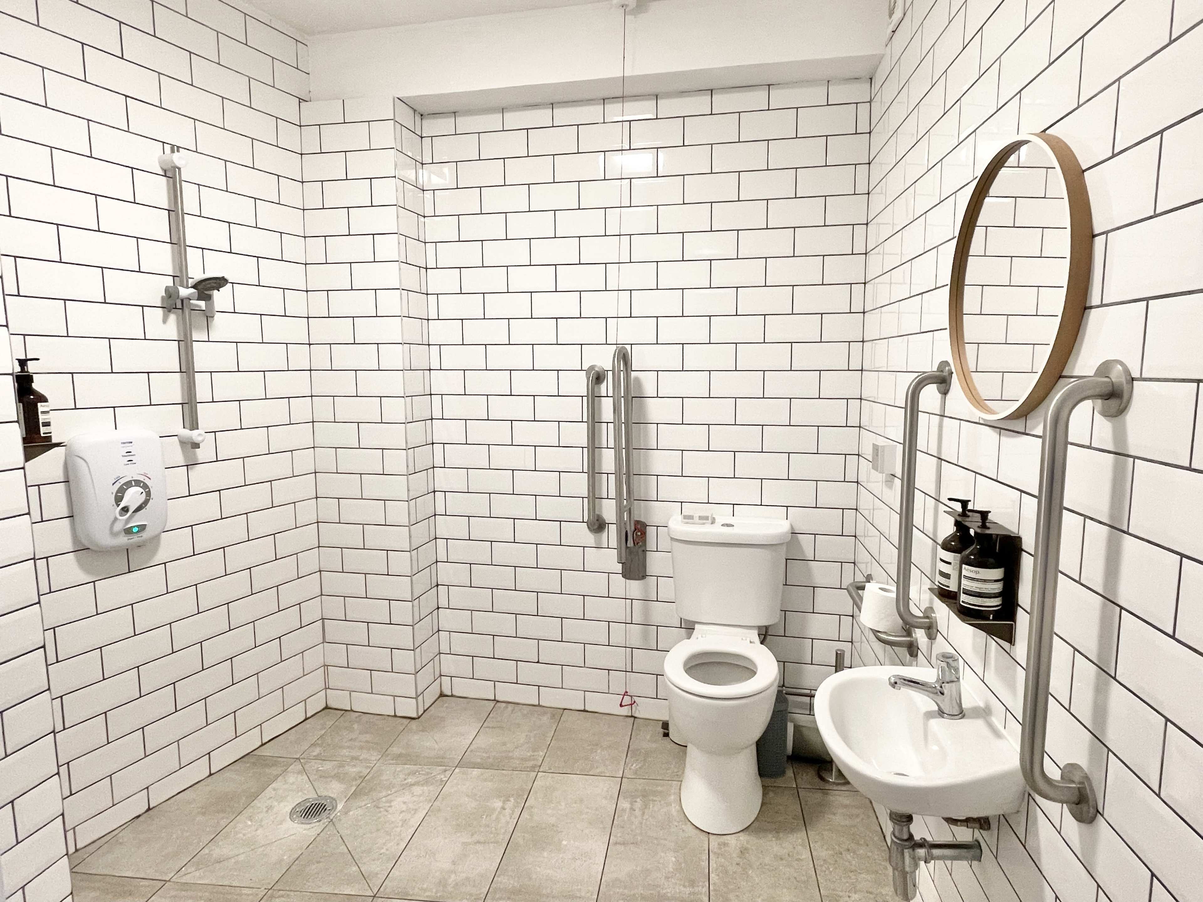 The image shows a clean, modern bathroom with white subway tile walls, a toilet, a shower area with grab bars, and a circular mirror.