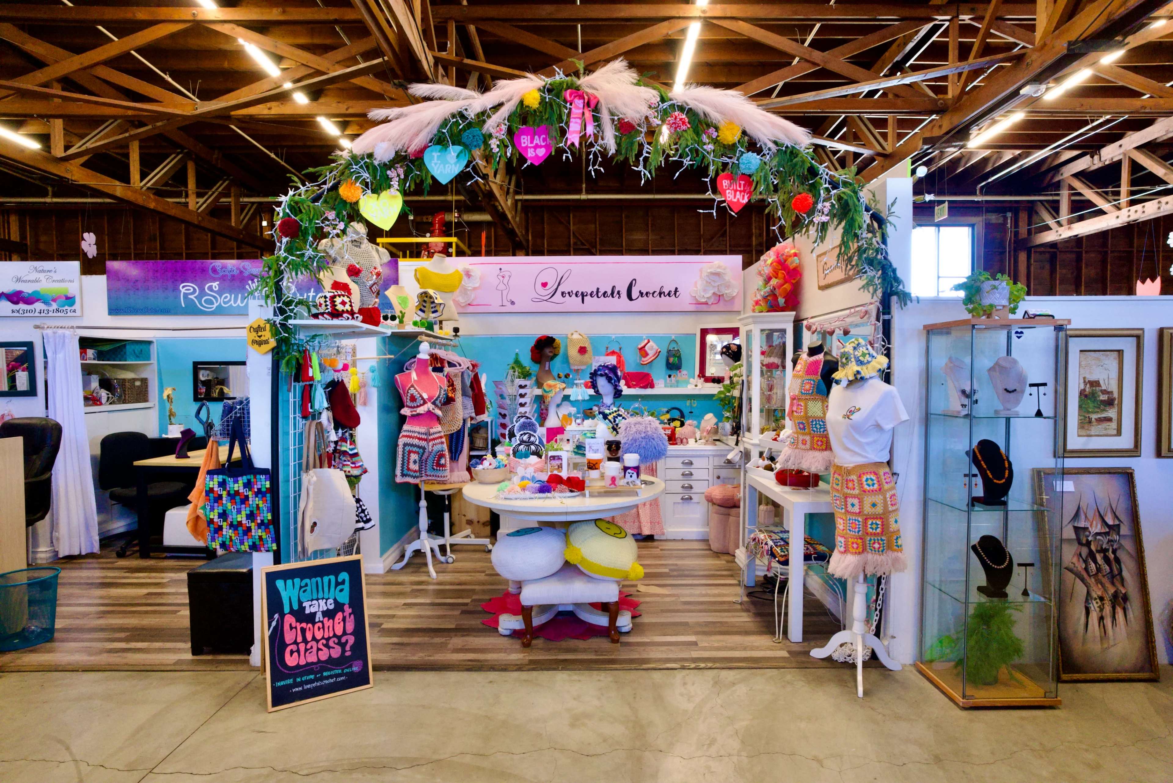 A colorful booth features a variety of handmade clothing and accessories, adorned with a floral arch, in a lively market setting.