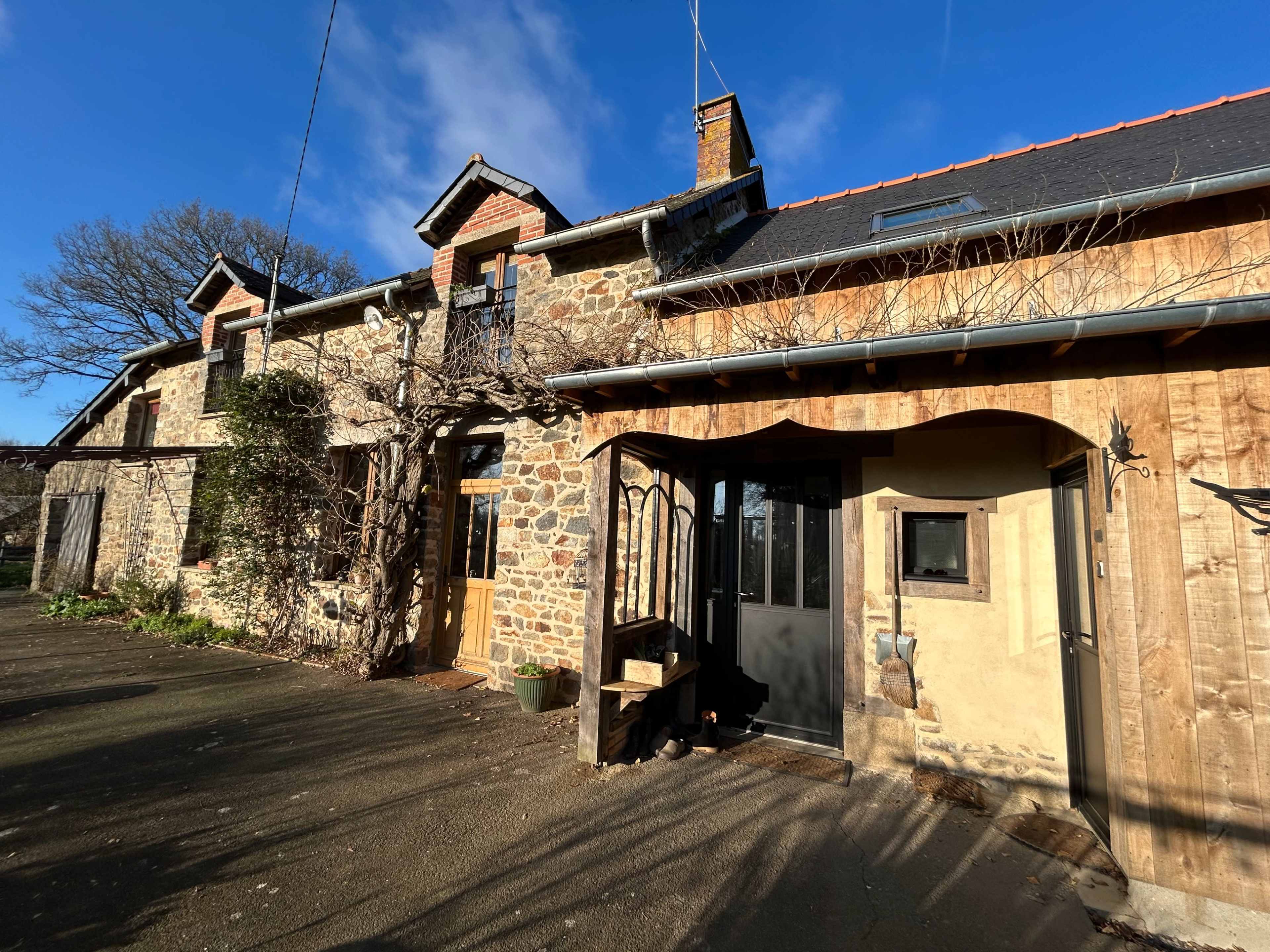 The image shows a rustic stone house with a wooden extension and a clear blue sky overhead.