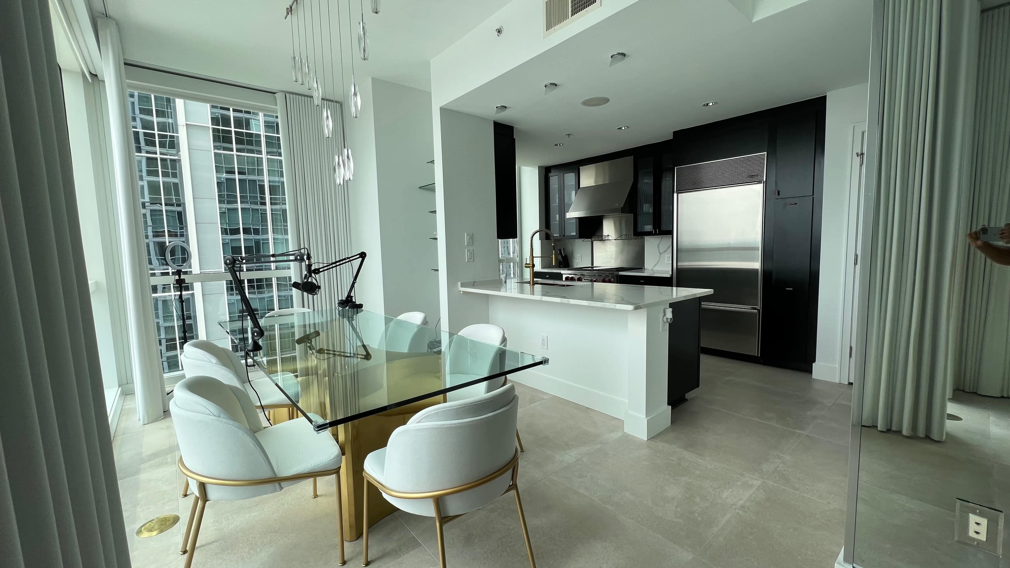The image shows a modern dining area with a glass table and white chairs, adjacent to a sleek kitchen featuring stainless steel appliances.