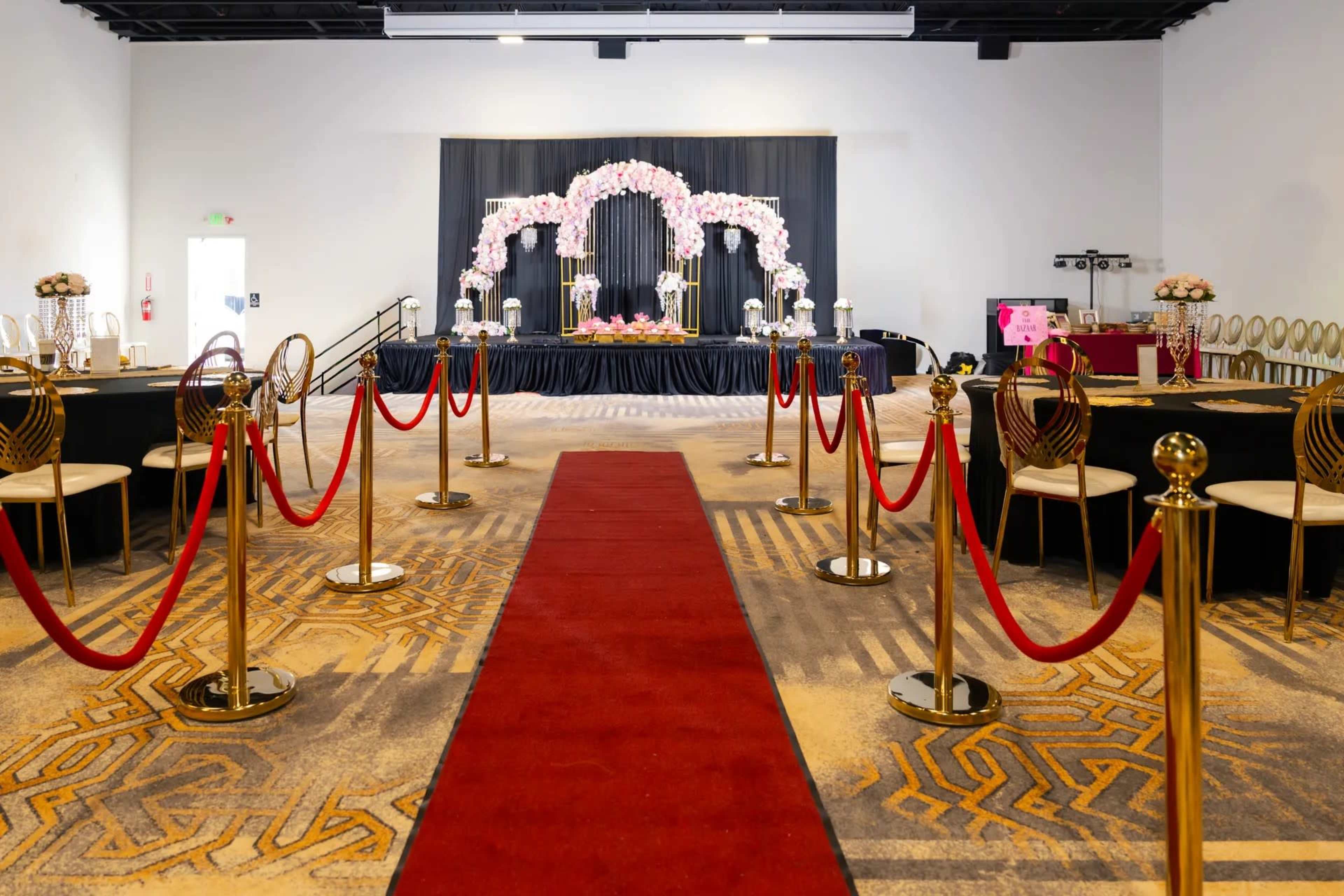 The image shows a banquet hall set up with a red carpet leading to a decorated stage featuring floral arrangements and tables arranged on either side.