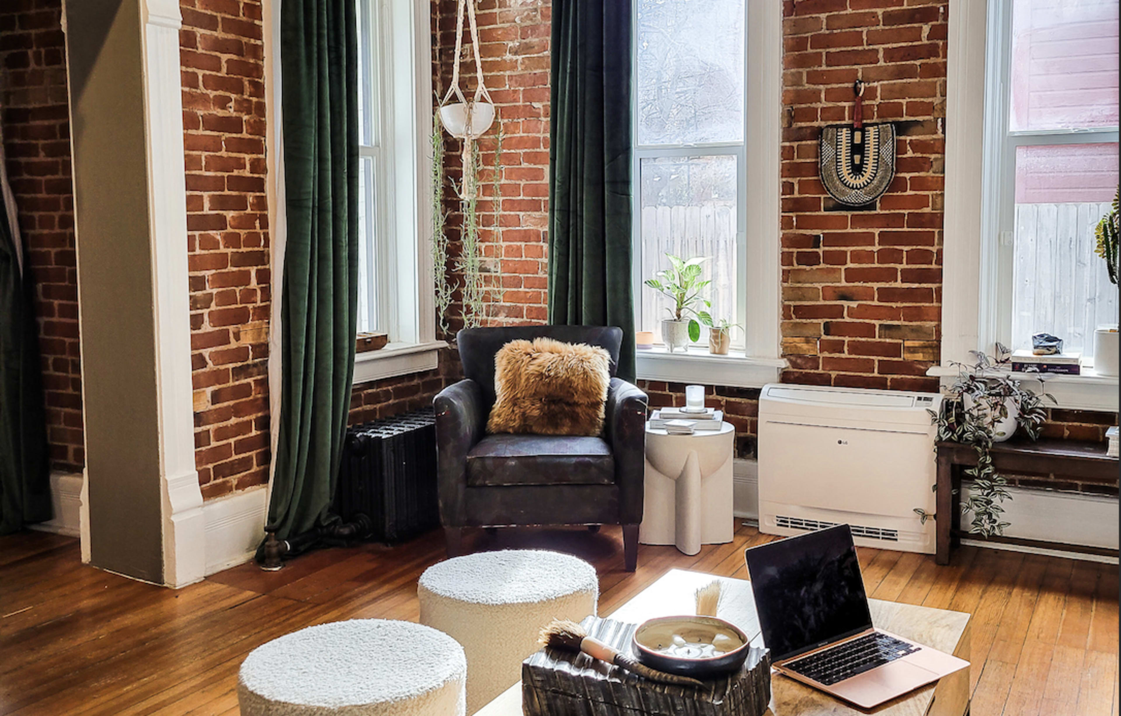 A cozy living room features a dark armchair with a fur throw, two round ottomans, a laptop on a wooden coffee table, and large windows framed by green curtains, all against a backdrop of exposed brick walls.