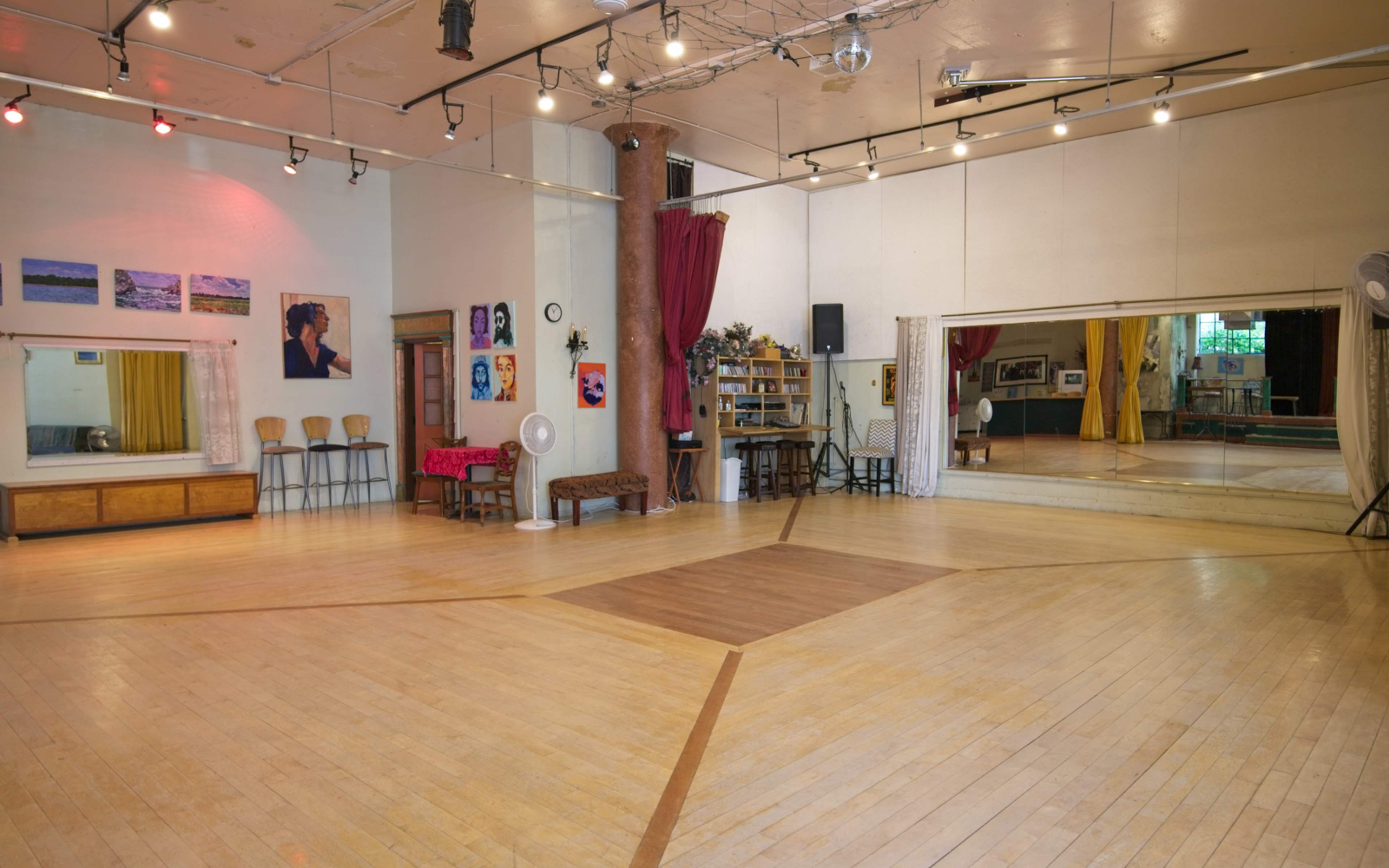 A spacious dance studio with polished wooden floors, wall-mounted mirrors, and various decorations, including artwork and seating along the sides.