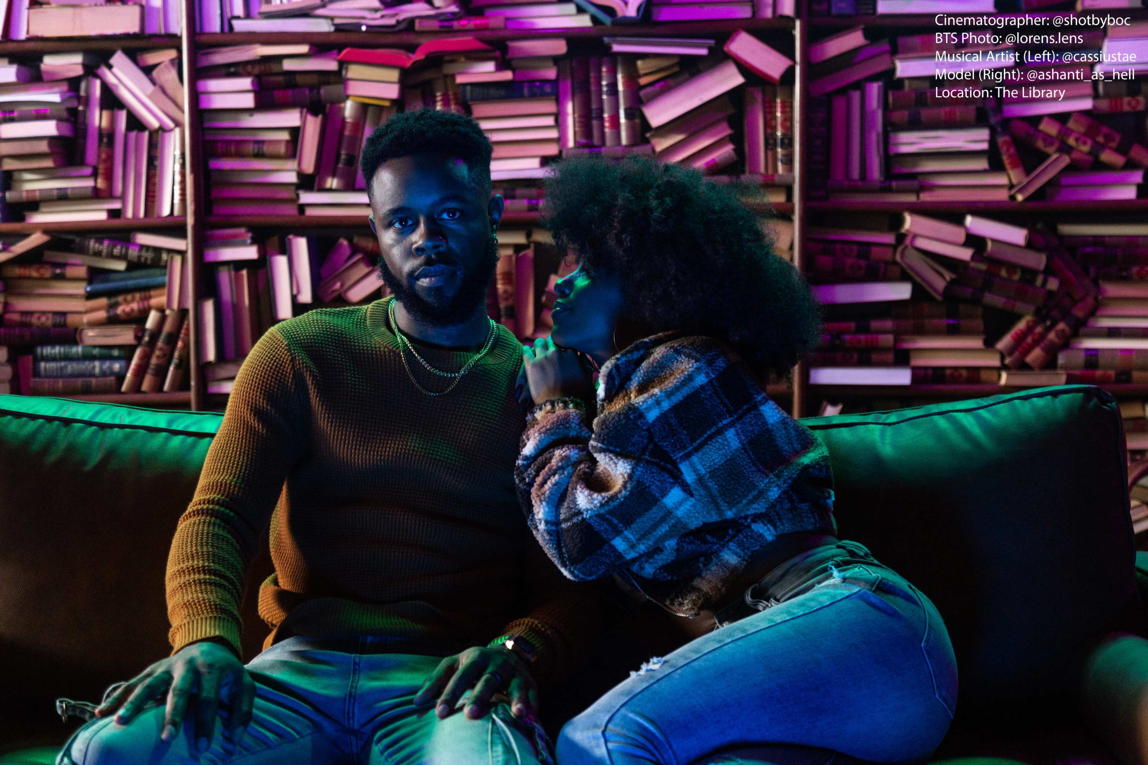 A man and a woman sit closely together on a couch in a dimly lit library filled with books.