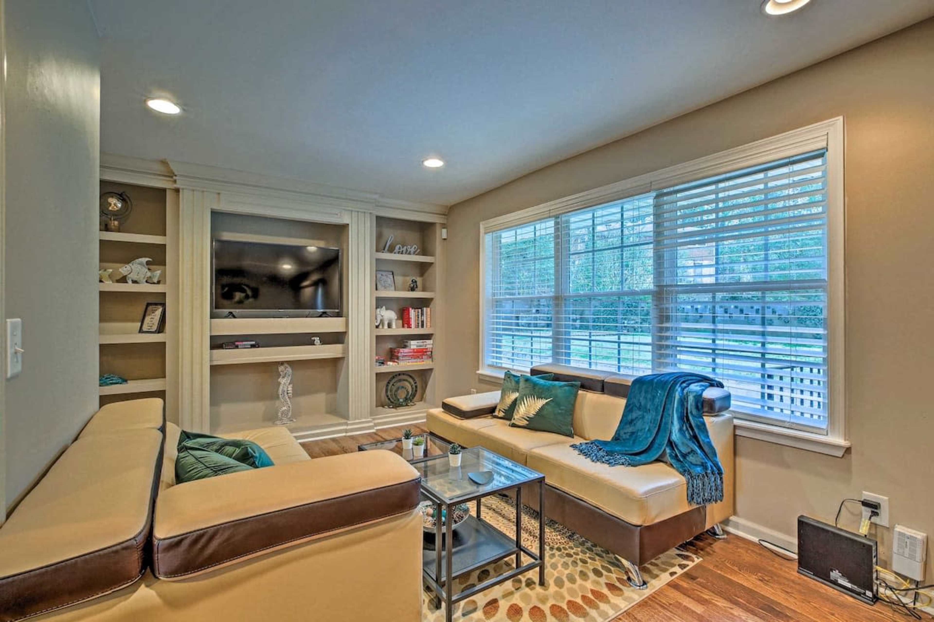 A living room with a beige sofa, a glass coffee table, built-in shelves, and large windows with white blinds.