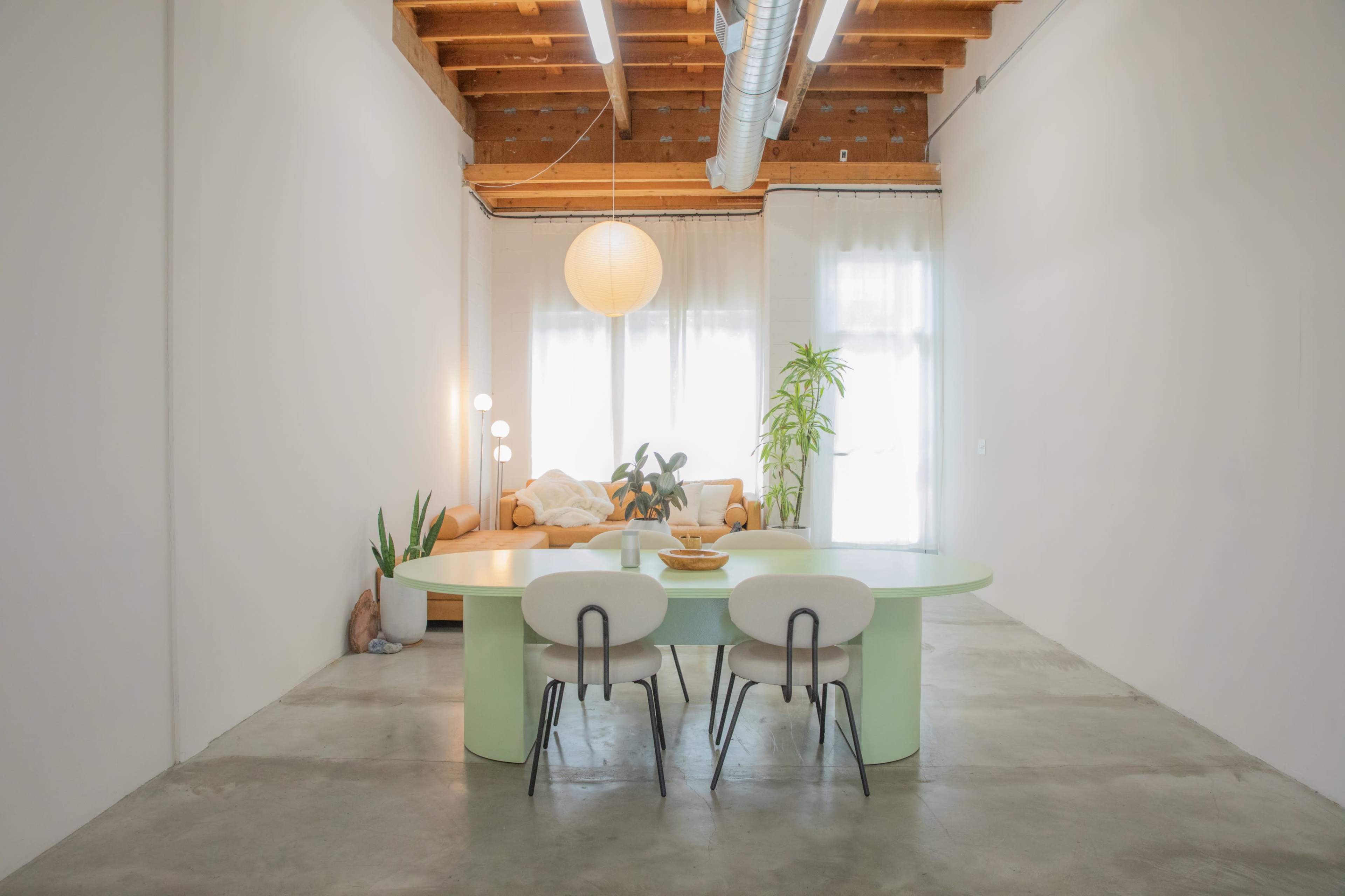 A minimalist room with a round green dining table, three chairs, a sofa in the background, and large windows letting in natural light.