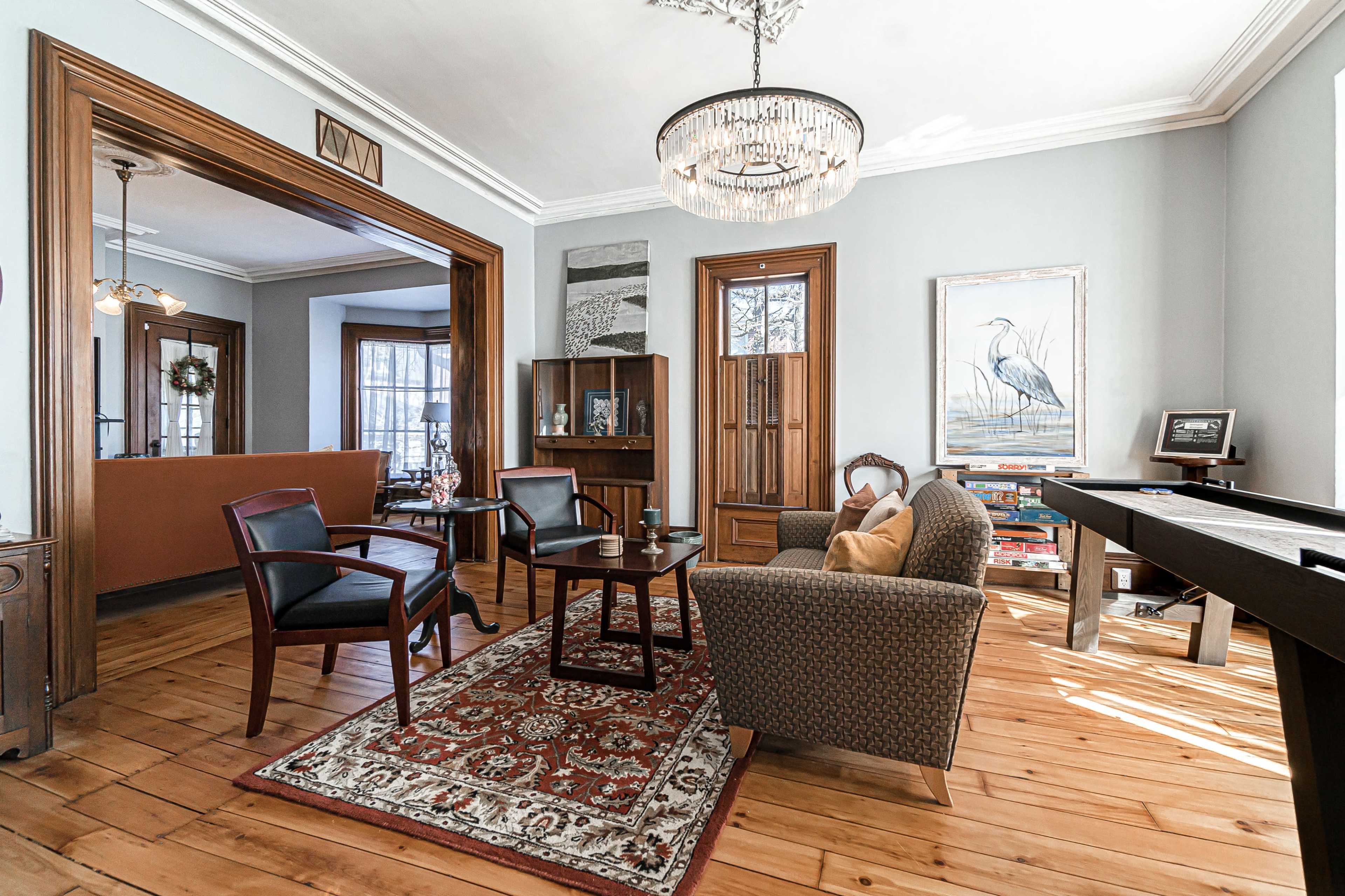 A well-lit living room with a mix of modern and traditional furnishings, featuring a patterned rug, two chairs, a small table, and a view into an adjoining room with bookshelves.