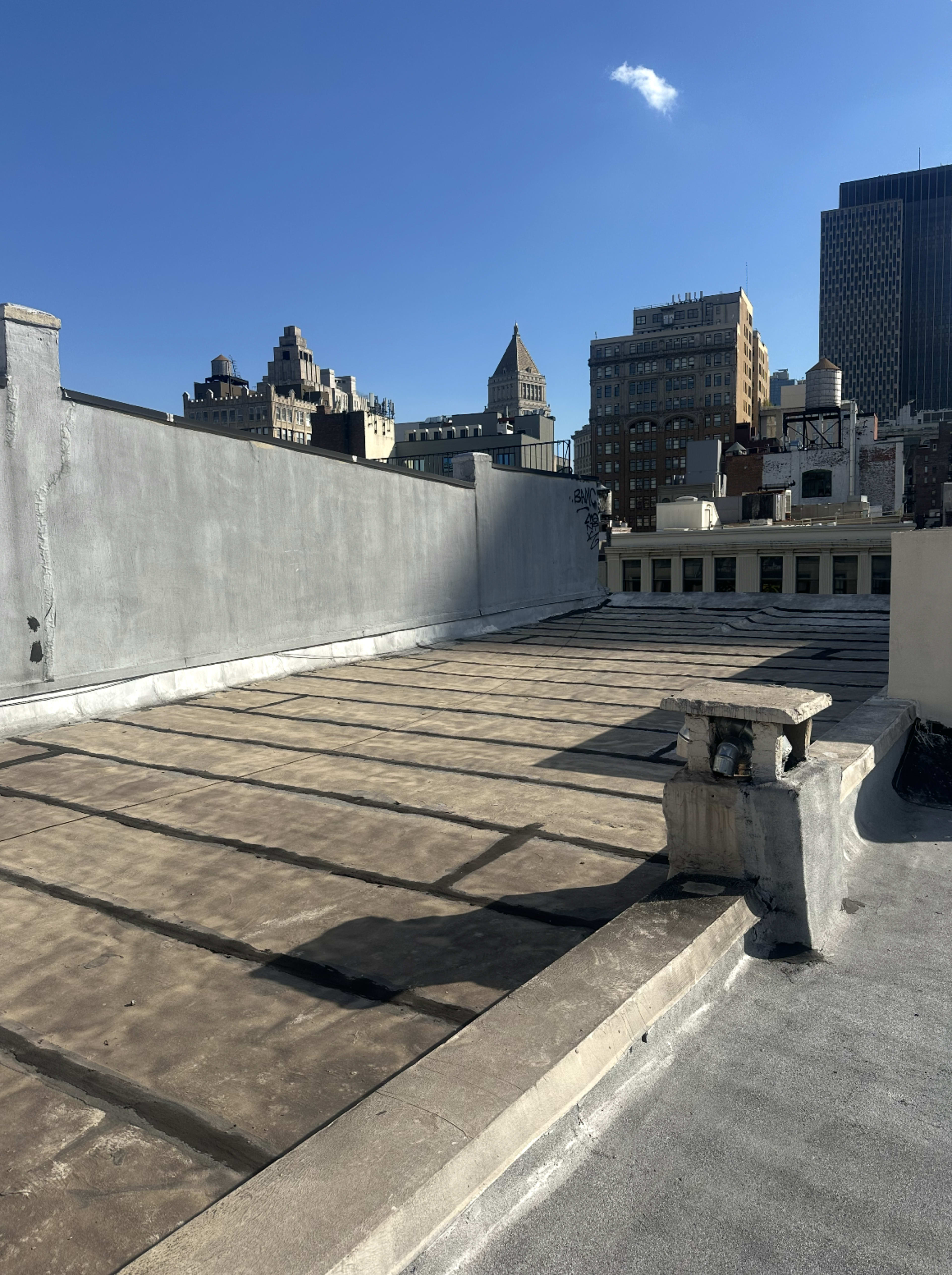 The image shows an empty rooftop with a flat, bare surface and a city skyline in the background under a clear blue sky.