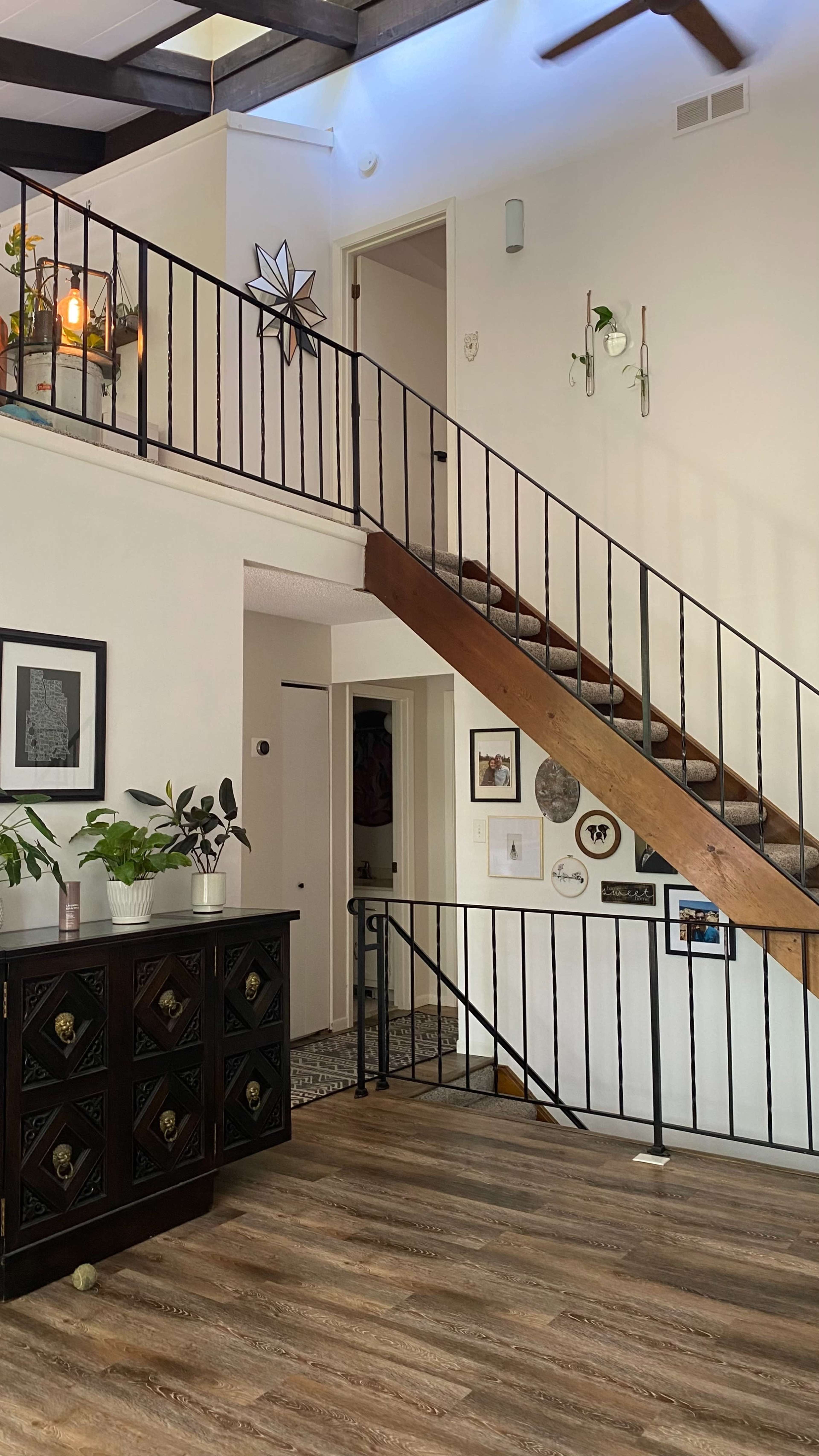 A two-story entryway featuring a wooden staircase, plants, wall art, and a dark cabinet against one wall.