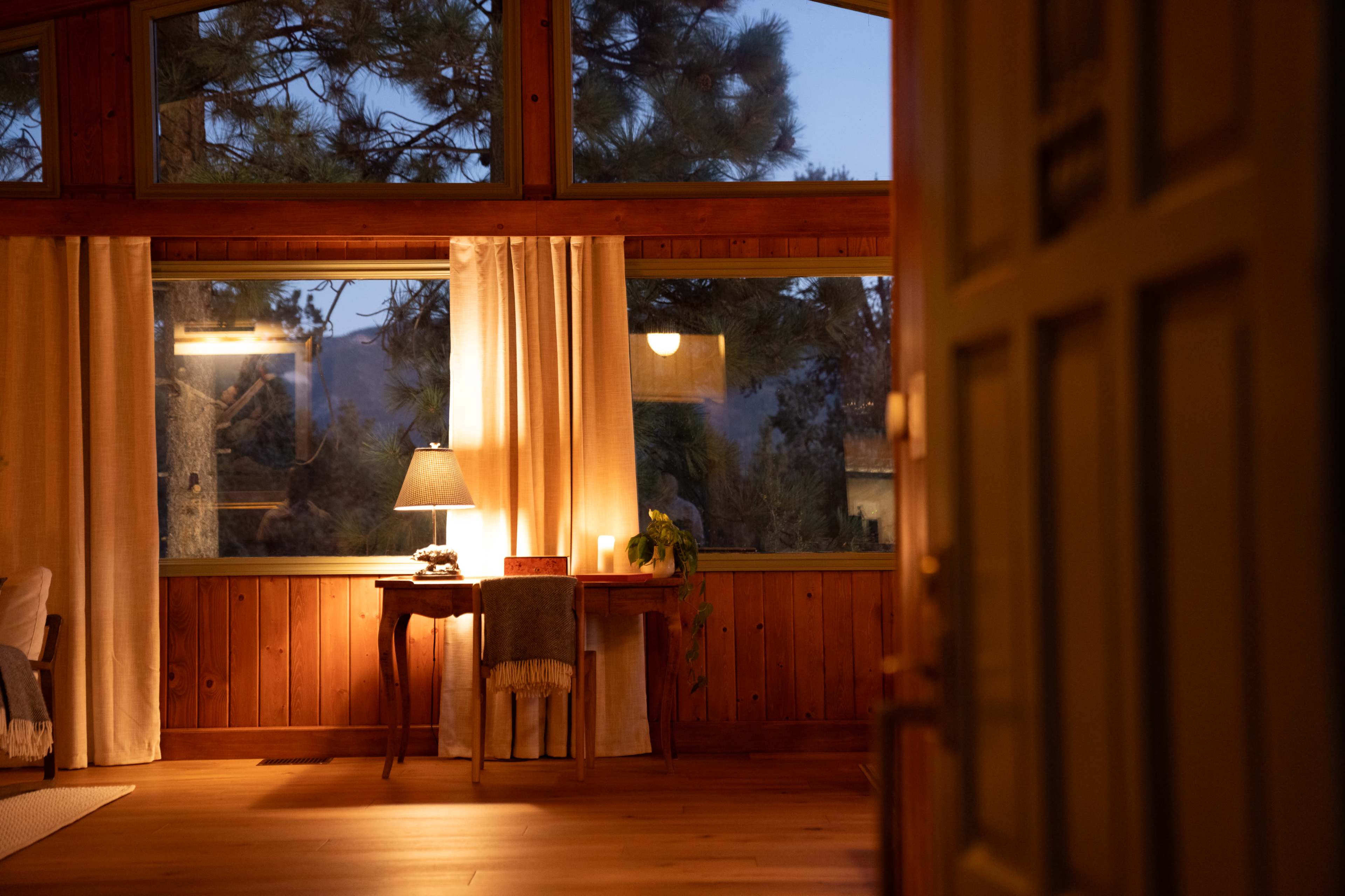 A warmly lit interior of a cabin features a wooden table with a lamp beside a large window displaying a nighttime view of trees.
