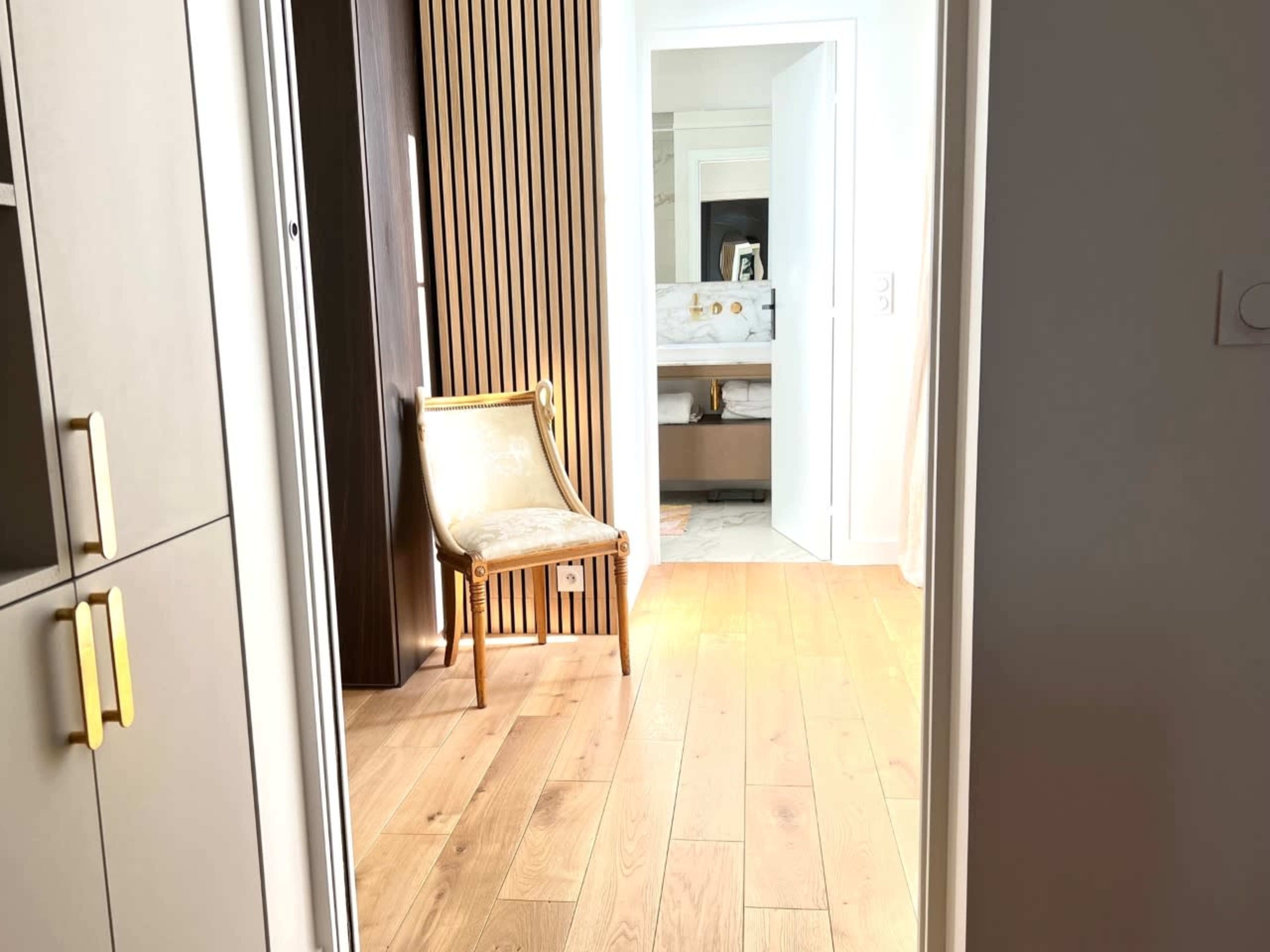 A hallway leading to a bathroom, featuring a wooden floor, a vintage chair, and a partially open door.