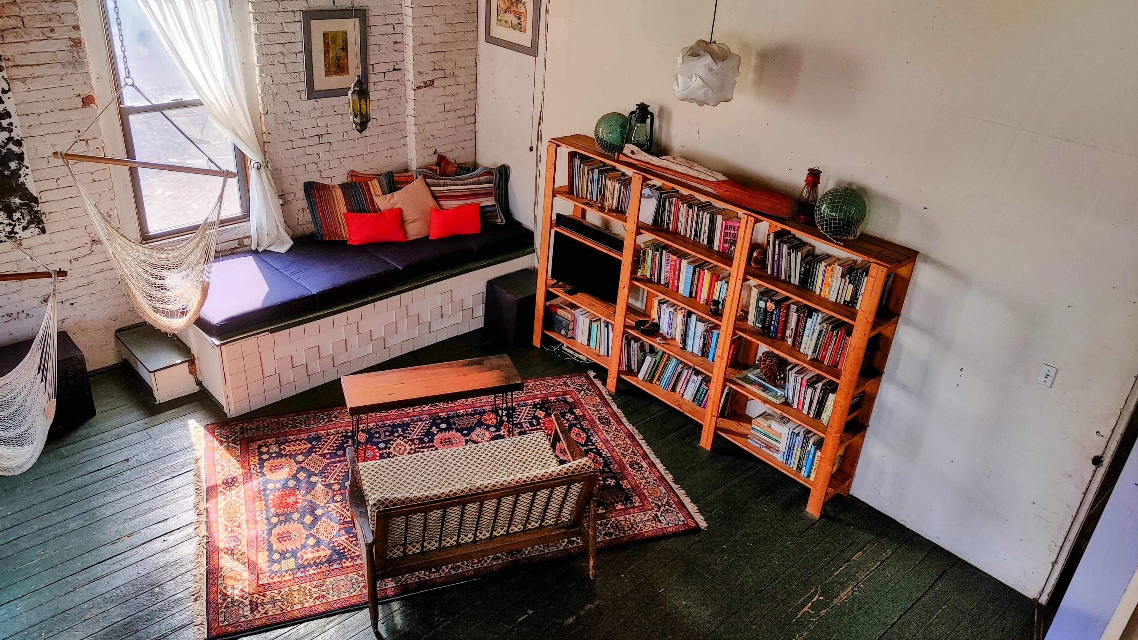 The image shows a cozy room featuring a seating area with a rug, a bookshelf filled with books, and a hammock by the window.