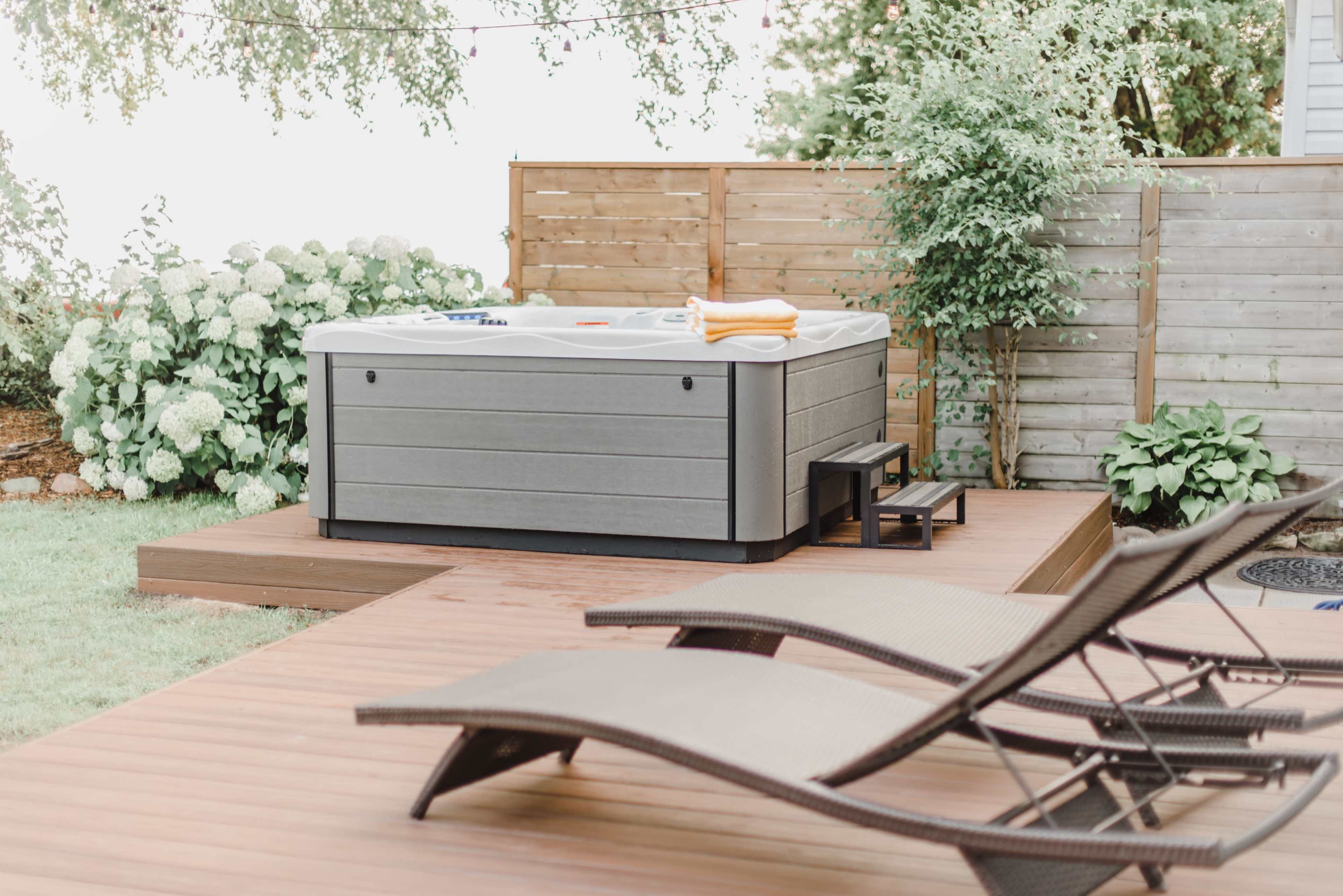 A hot tub sits on a wooden deck surrounded by greenery and lounge chairs.