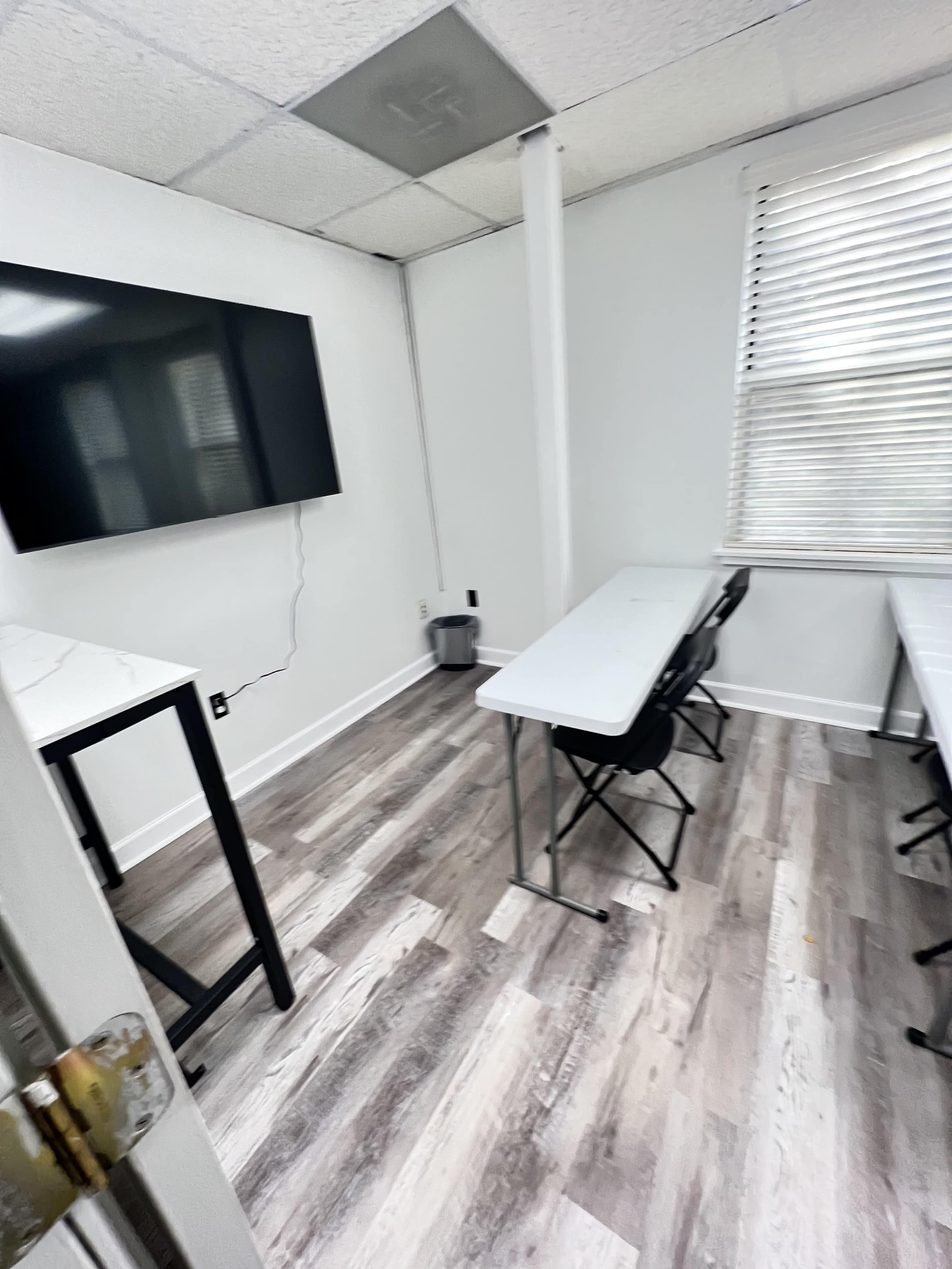 A small, bright classroom with a large wall-mounted television, a white table, and several black chairs arranged around it.
