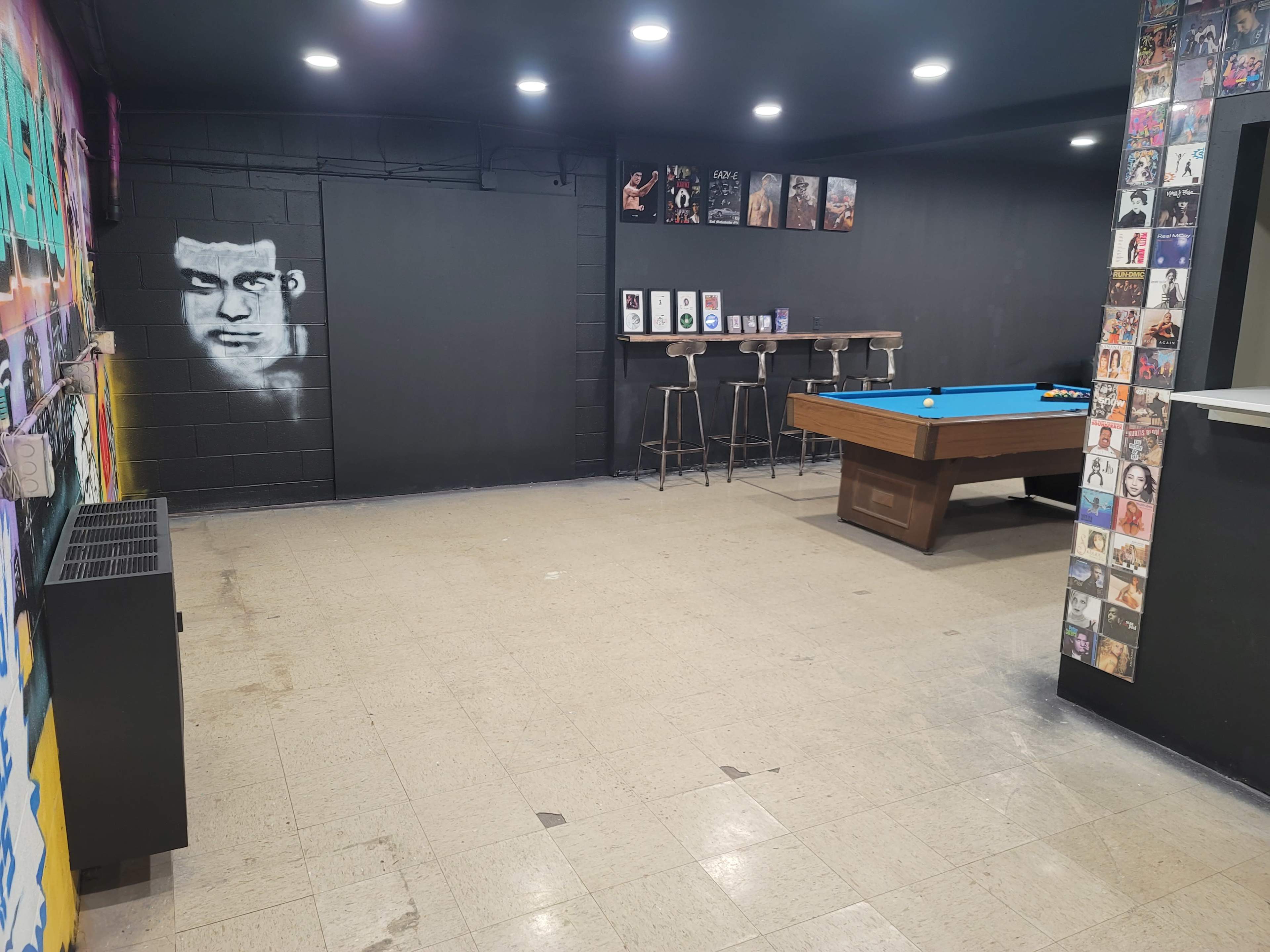 The image shows a dimly lit room with a pool table on one side and a wall covered in colorful artwork and framed pictures.