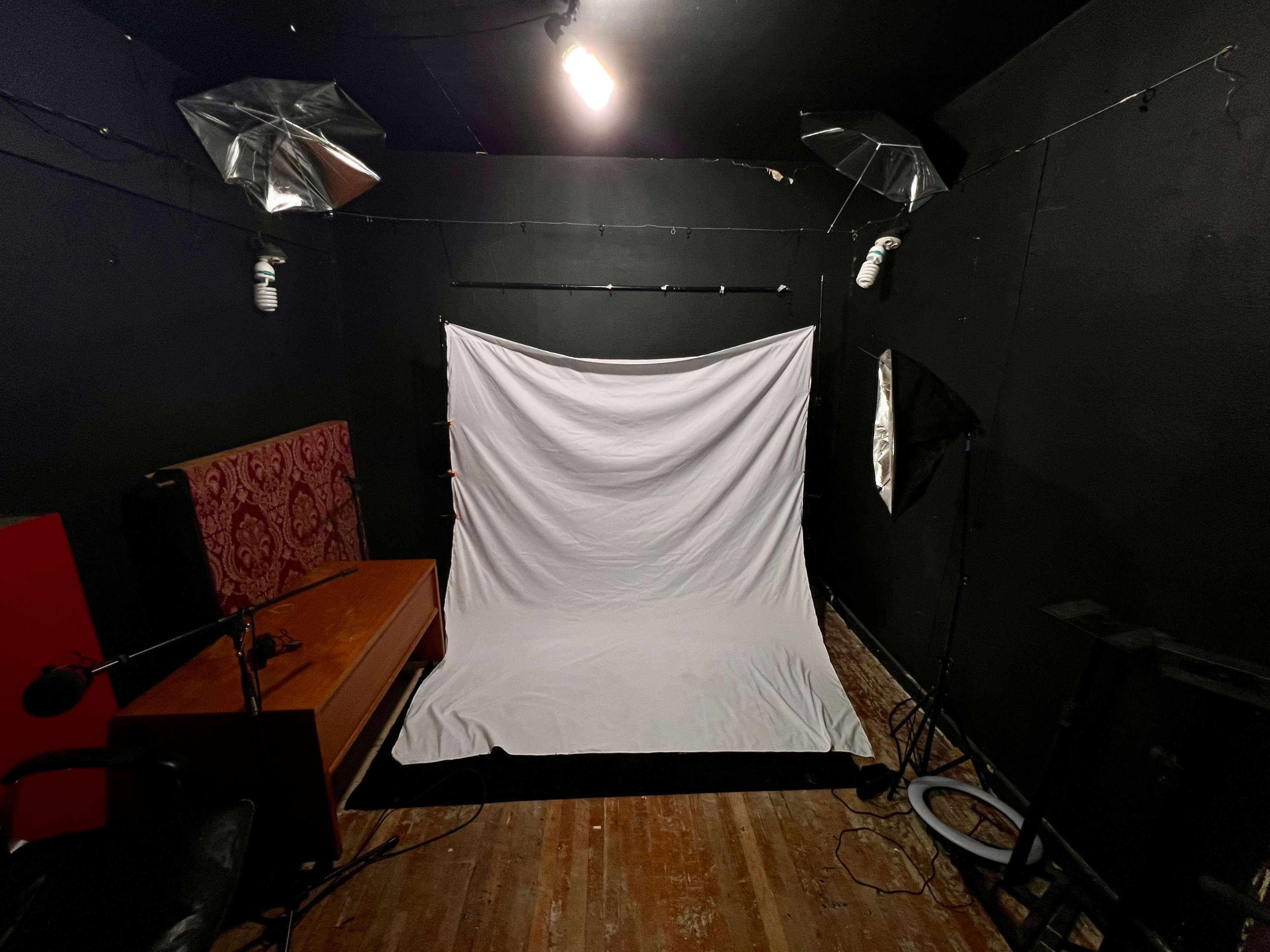 The image shows a small, dark studio space with a white backdrop, a wooden table, and lighting equipment positioned in the corners.