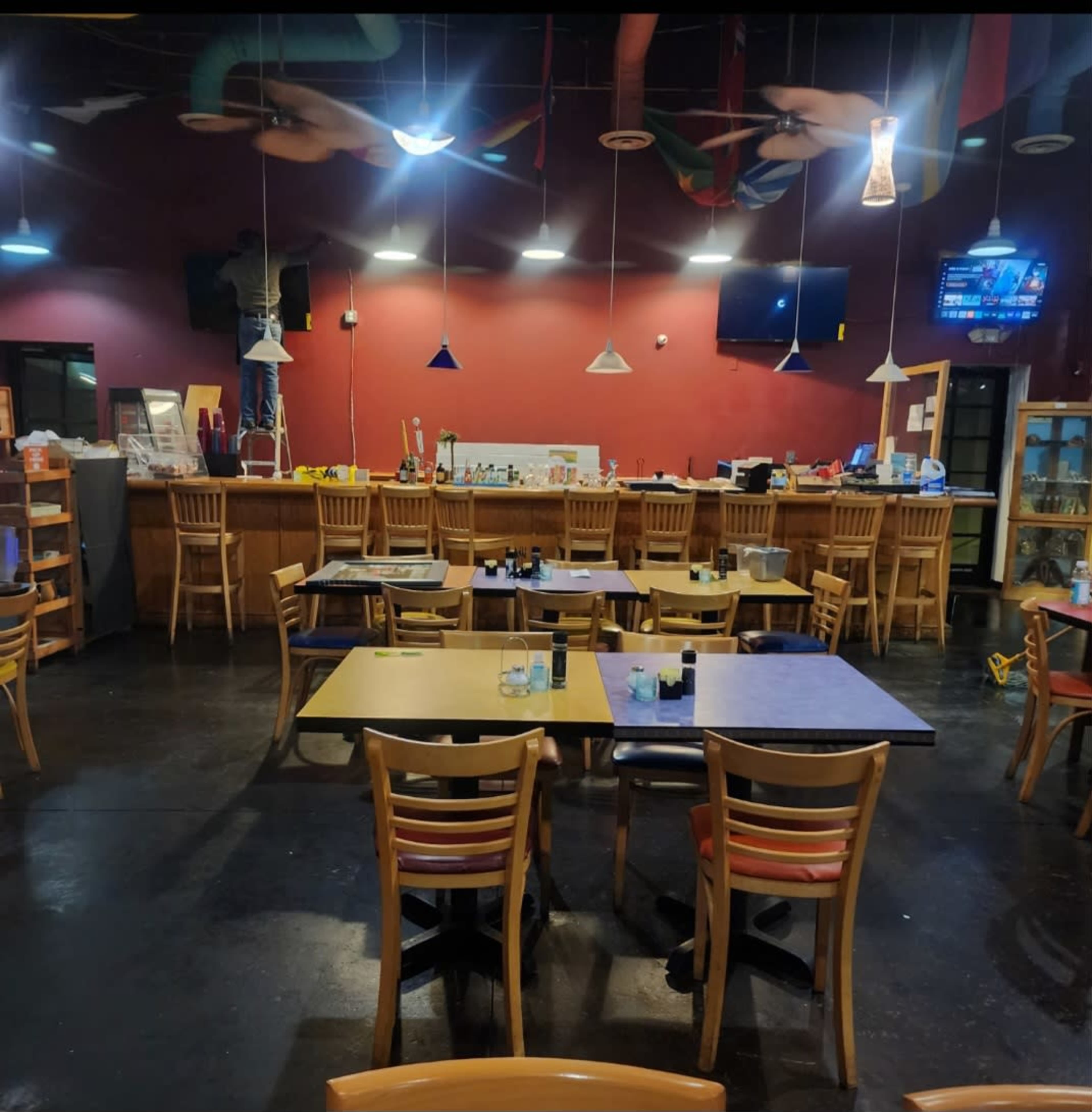 The image shows a partially empty restaurant with wooden tables and chairs, a bar in the background, and a person working on a ceiling repair.