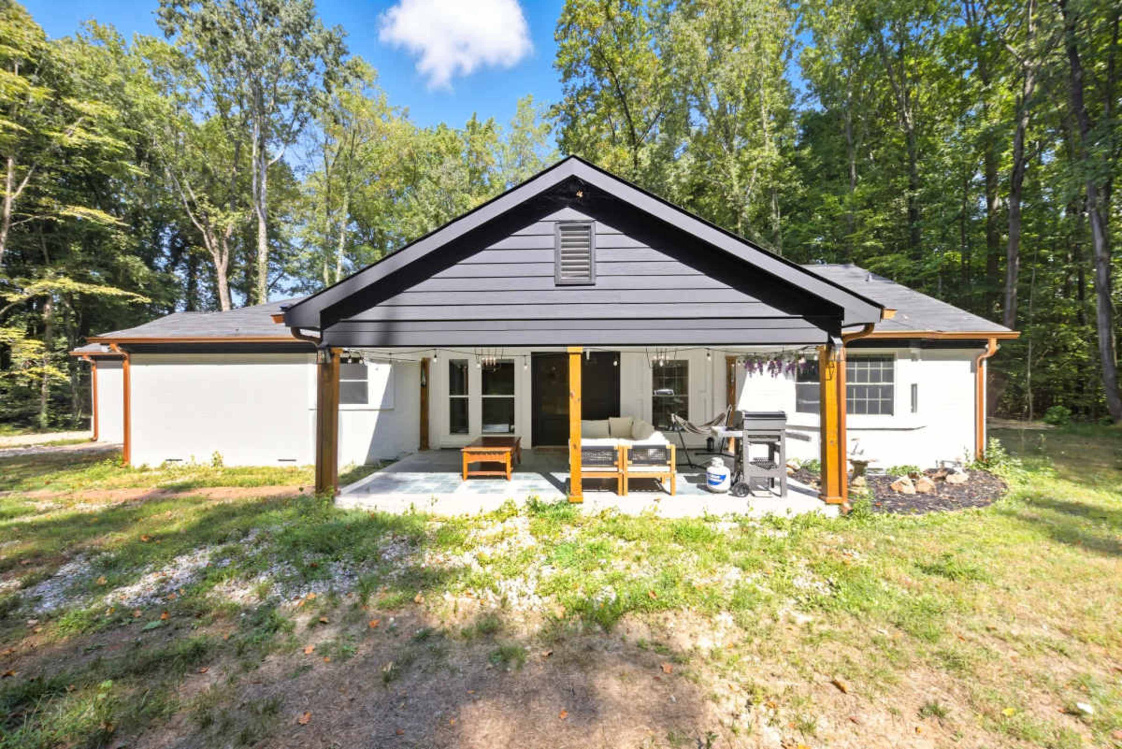 The image shows a single-story house with a dark roof and a porch, surrounded by greenery and trees.