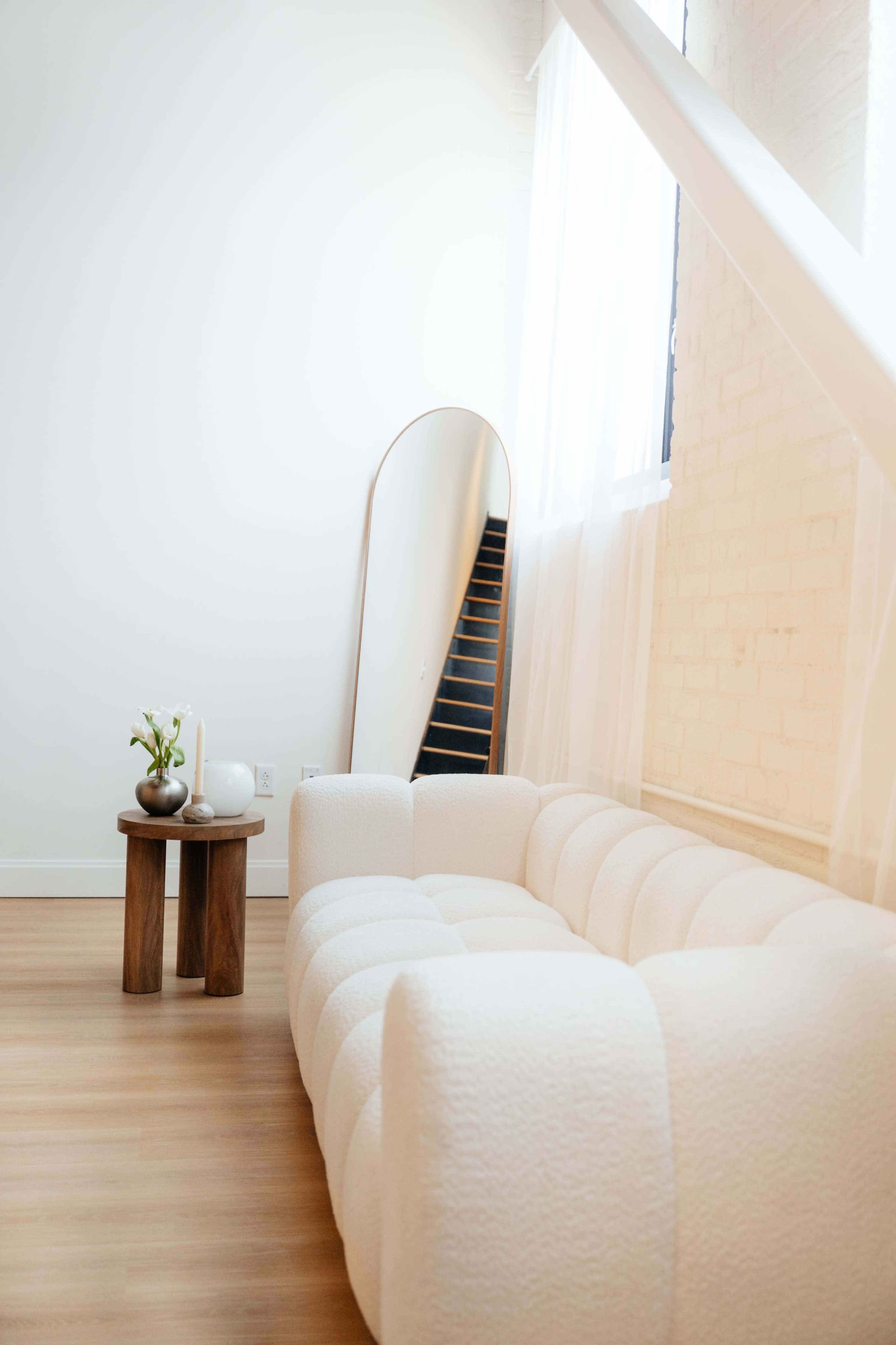 A light-colored couch is positioned next to a small wooden table in a room with a staircase and a large mirror reflecting natural light.