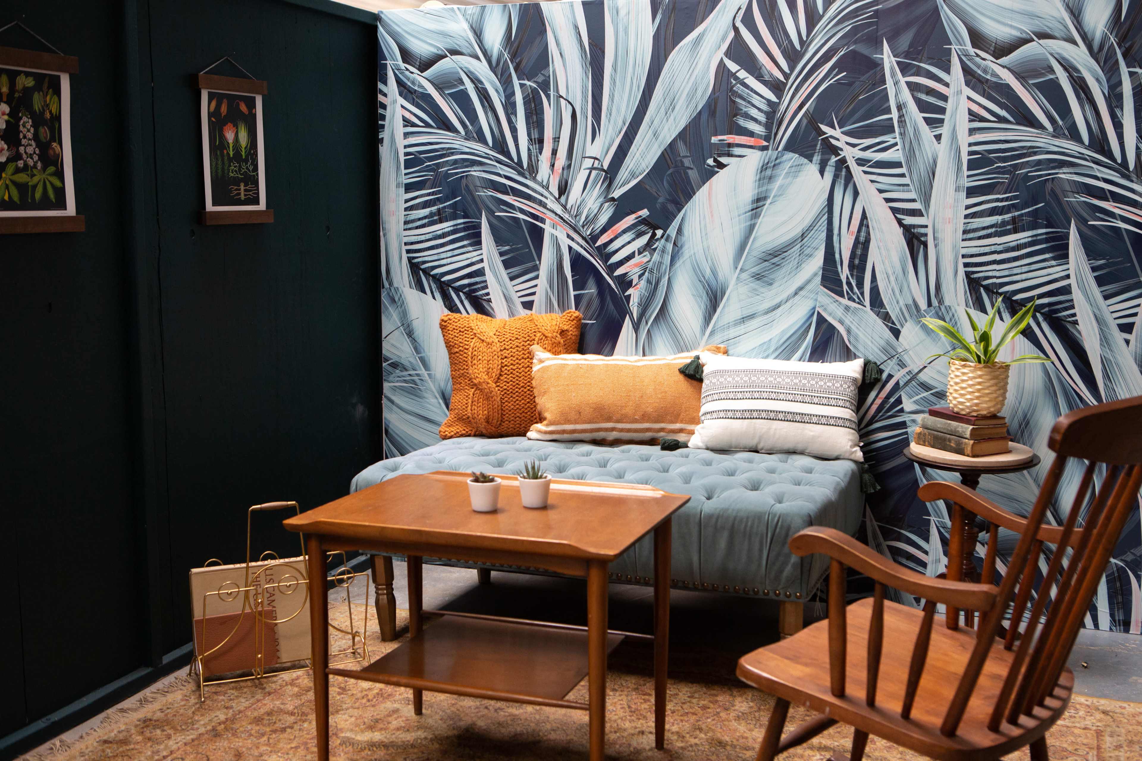 The scene features a cozy interior with a tufted blue couch, wooden coffee table, rocking chair, decorated wall with palm leaf prints, and potted plant.